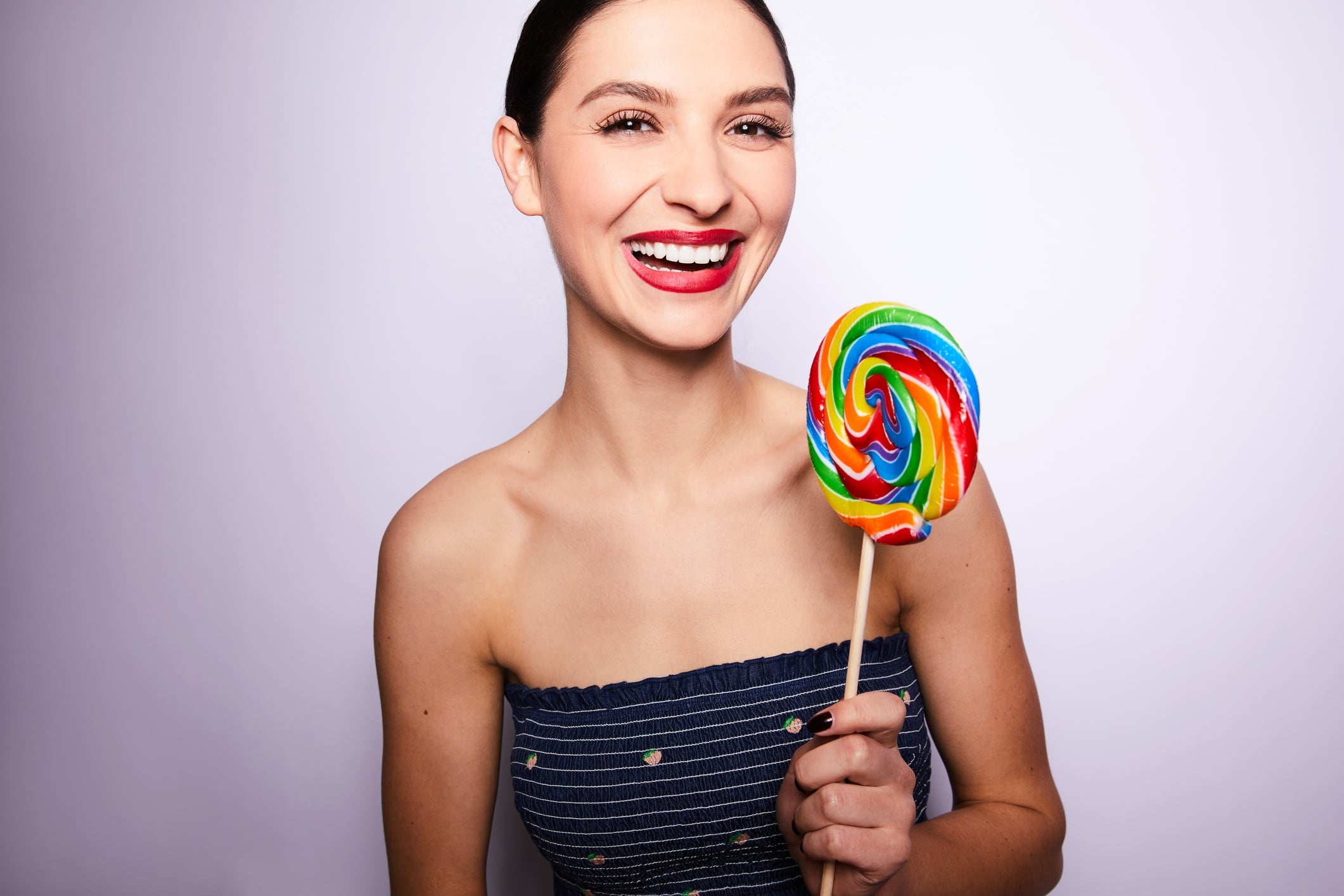 Studio portrait of a woman laughing and holding a large multicoloured lollipop. Purple background.