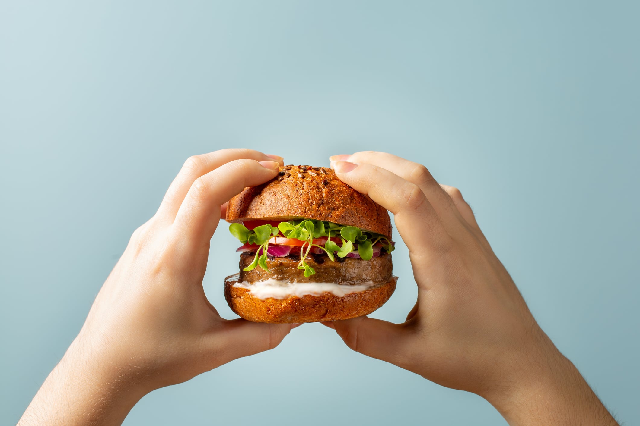 Hands holding burger. Hamburger with meat free plant based cutlet, tomatoes, onions, white sauce and microgreens on blue background. Healthy vegan or vegetarian food concept.