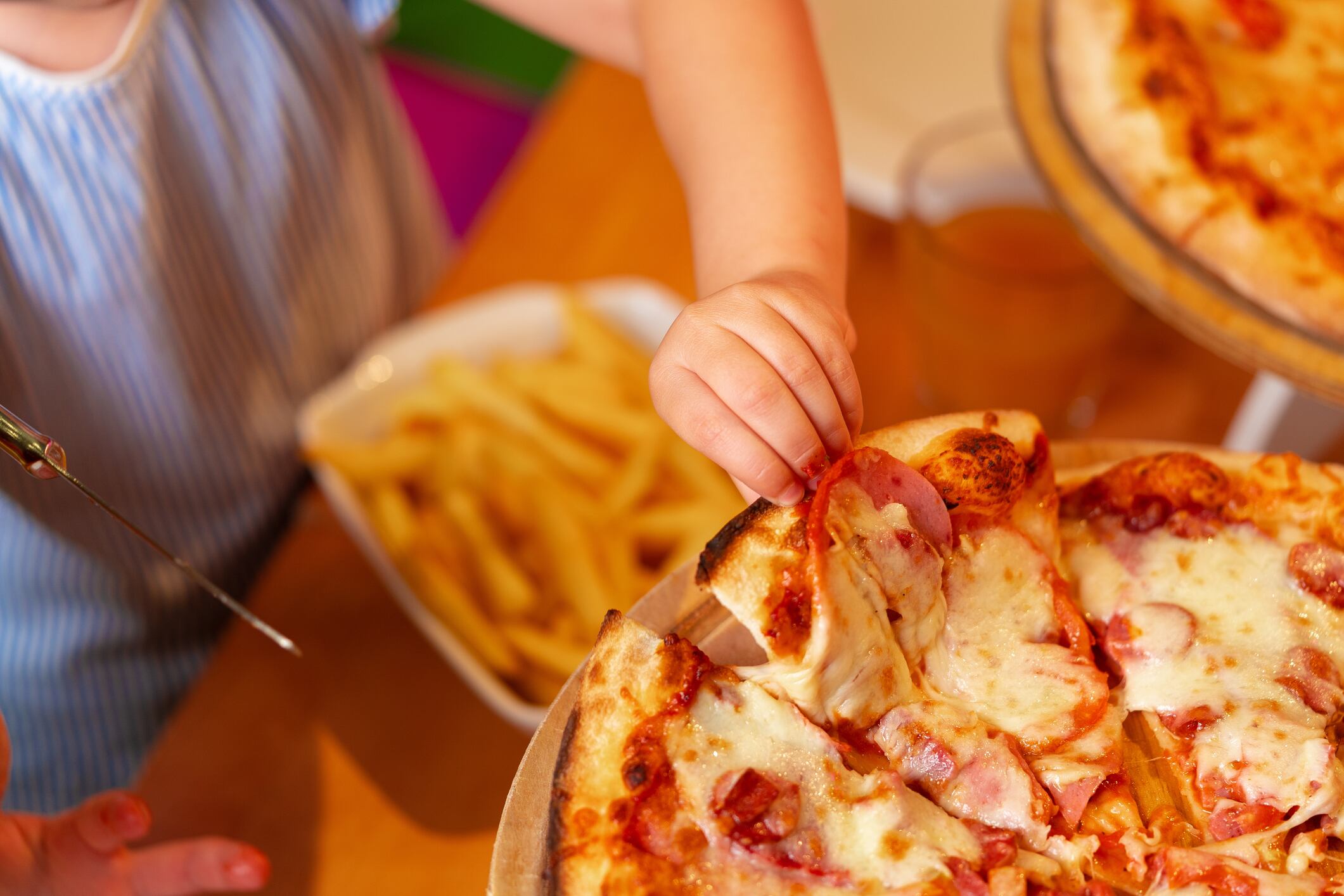 A young girl with light hair is captured in the moment of eating a slice of cheesy pizza, while another child is seated next to her, slightly out of focus. The scene suggests a casual birthday party setting with children enjoying food.