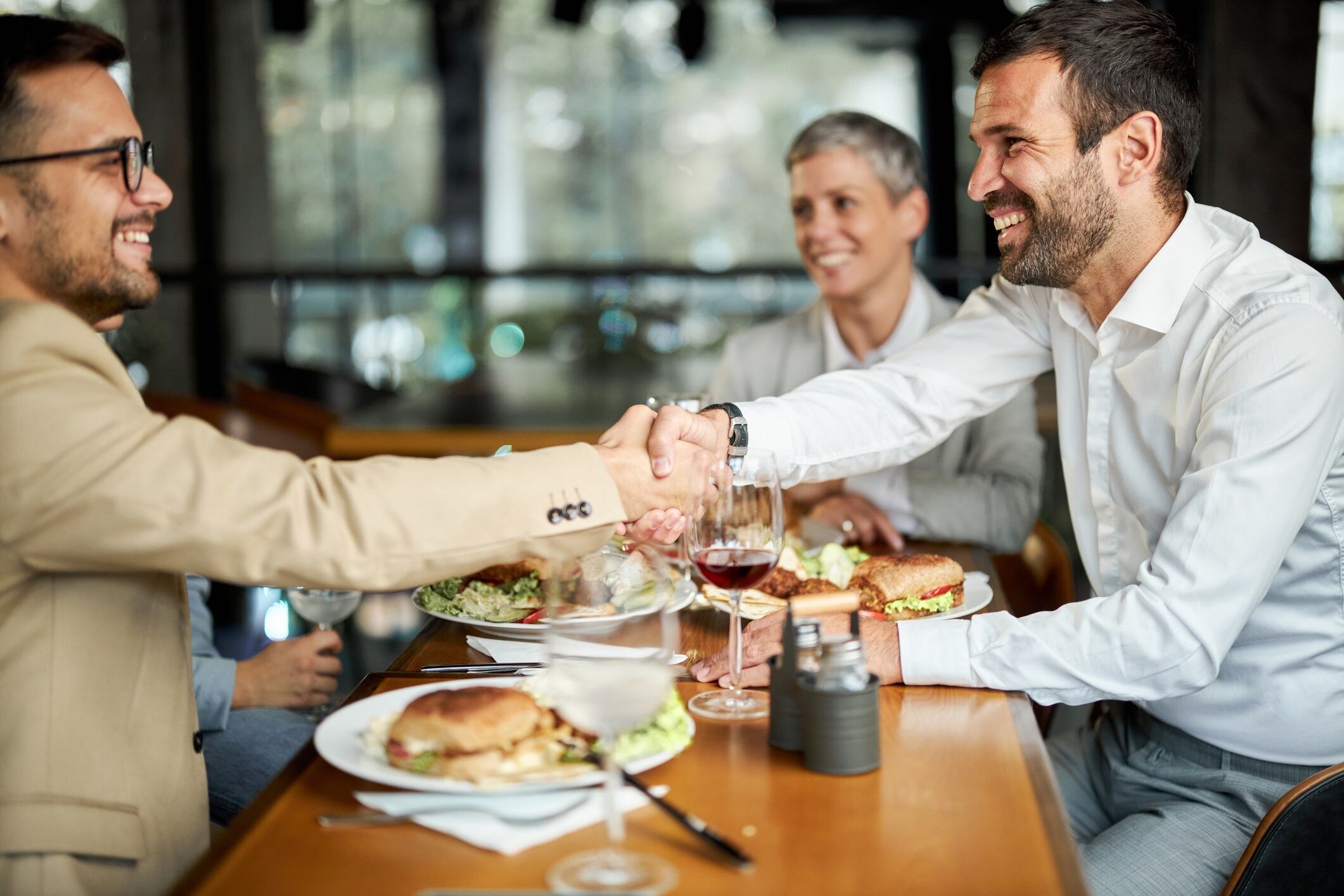 Happy businessmen shaking hands while having a lunch with their females colleague in a restaurant.