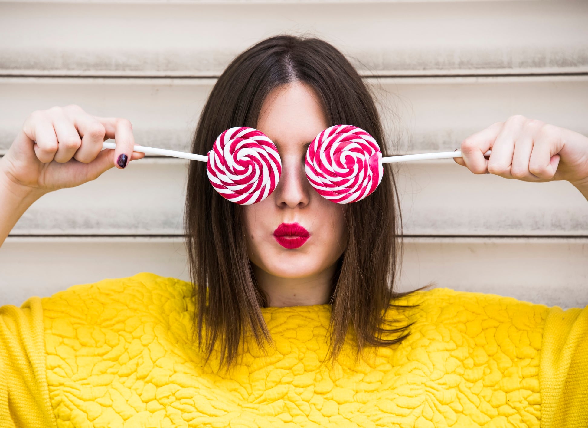 Brunette woman holding lollipops over both eyes. Red lipstick and bright yellow top.