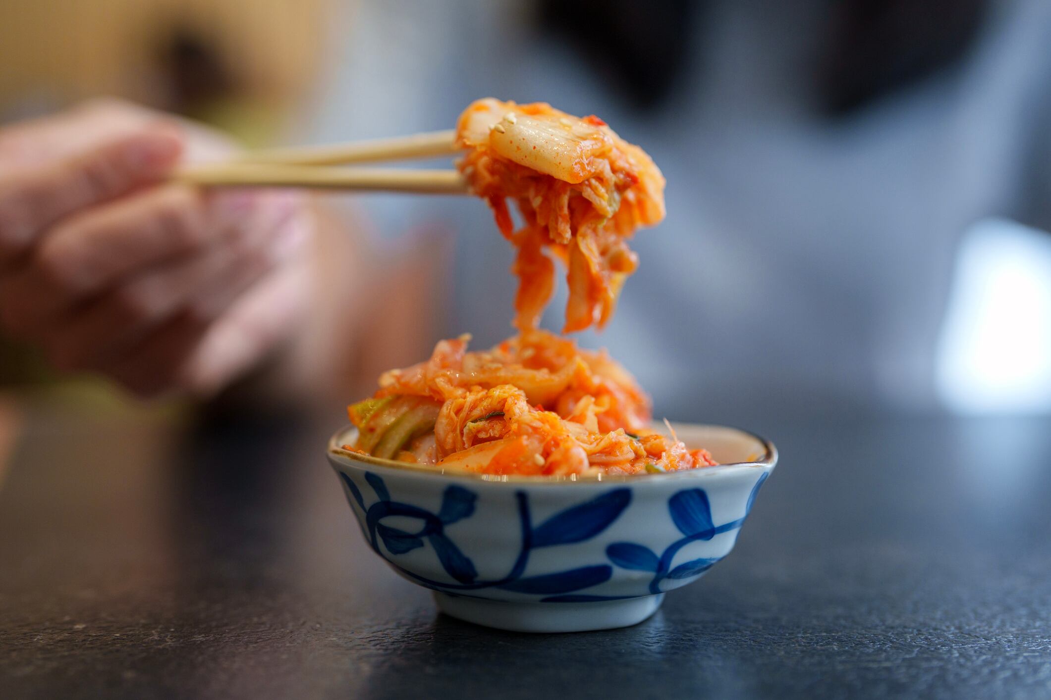 Close-up of a woman hand picking up fresh Korean kimchi from a patterned bowl using chopsticks, highlighting traditional Korean cuisine.