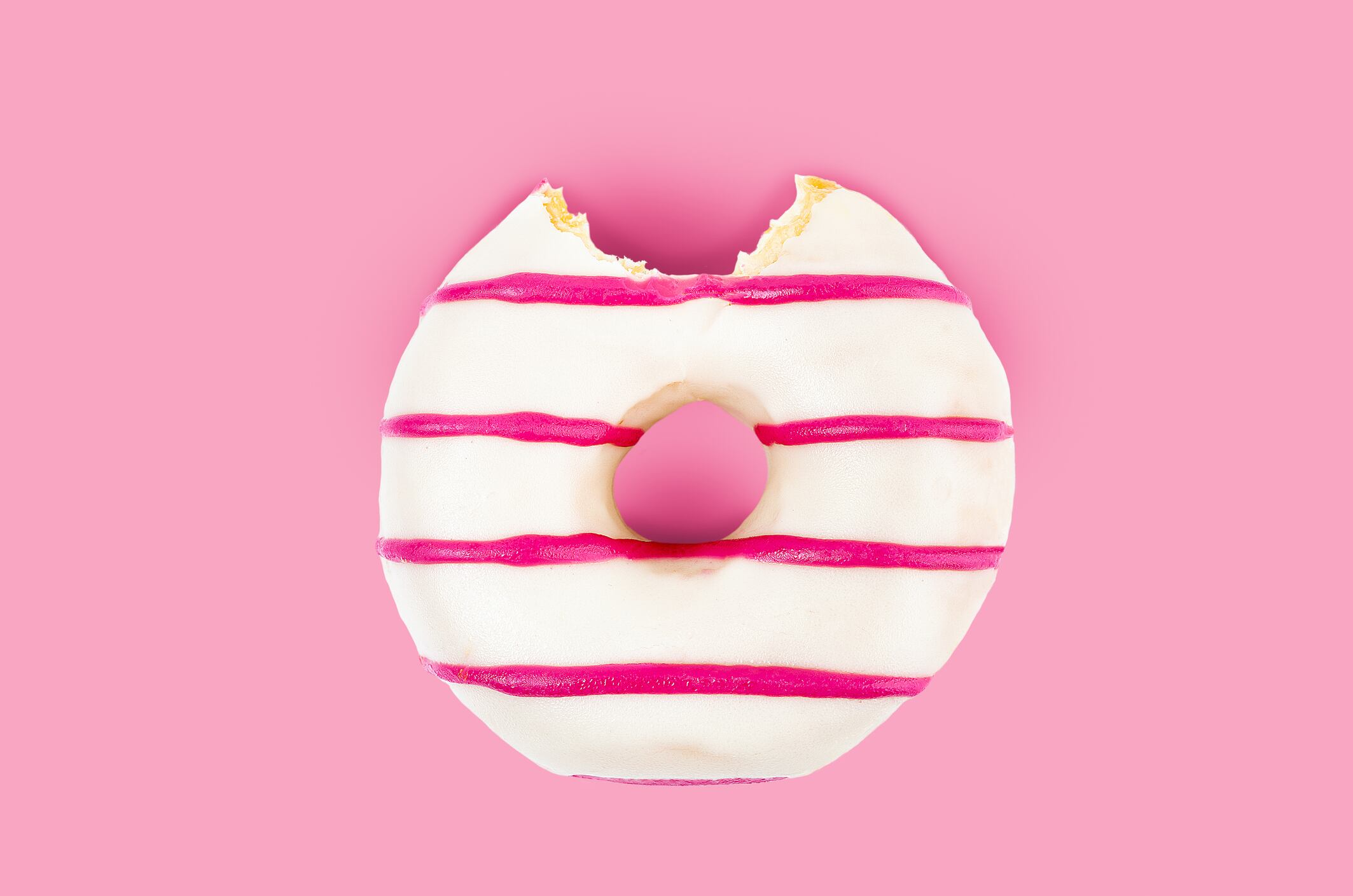 One bite missing of pink striped and white chocolate donut on pink background.