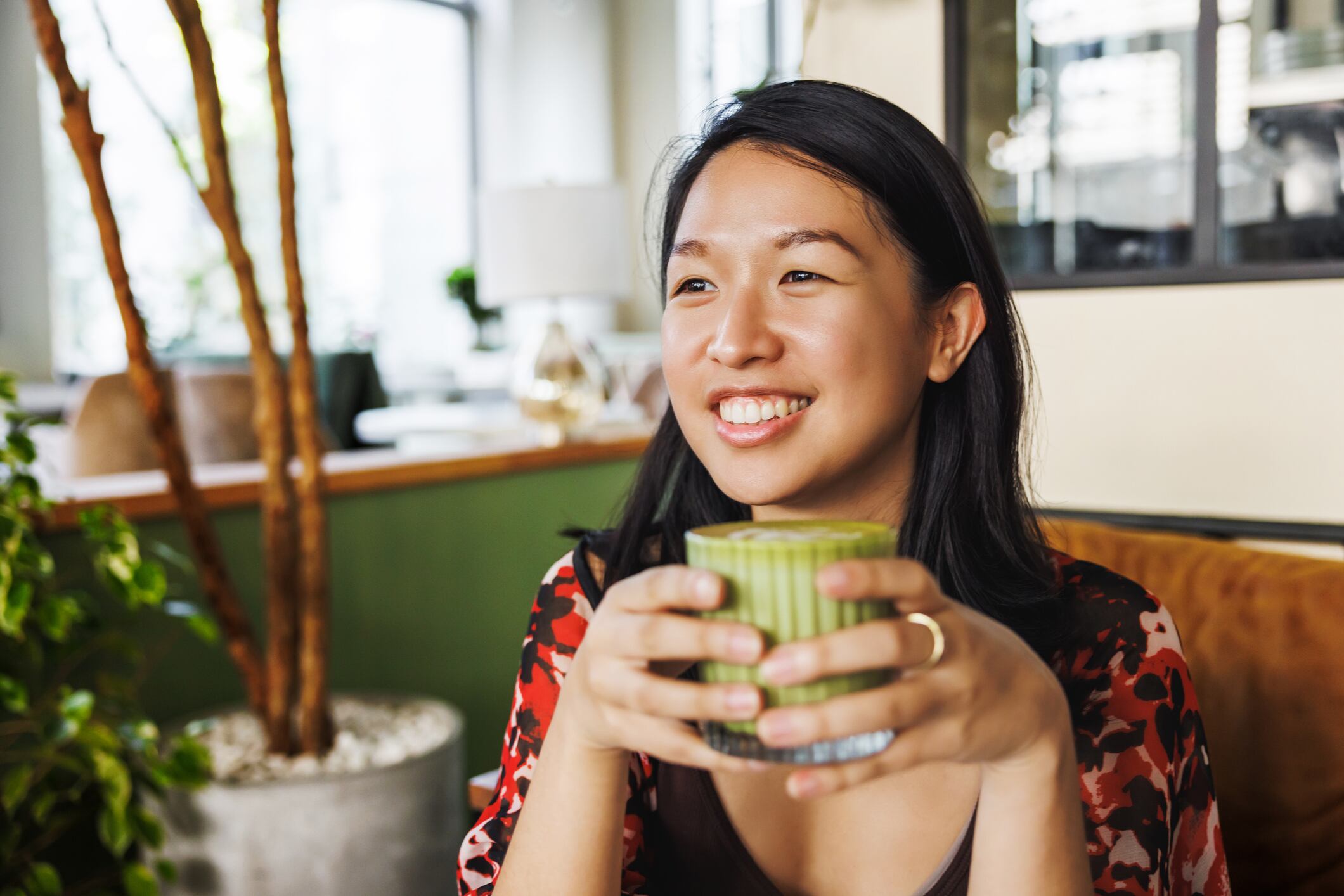 A cheerful woman holds a ceramic mug of matcha latte, her infectious smile enhancing the cozy and friendly vibe of the café around her.