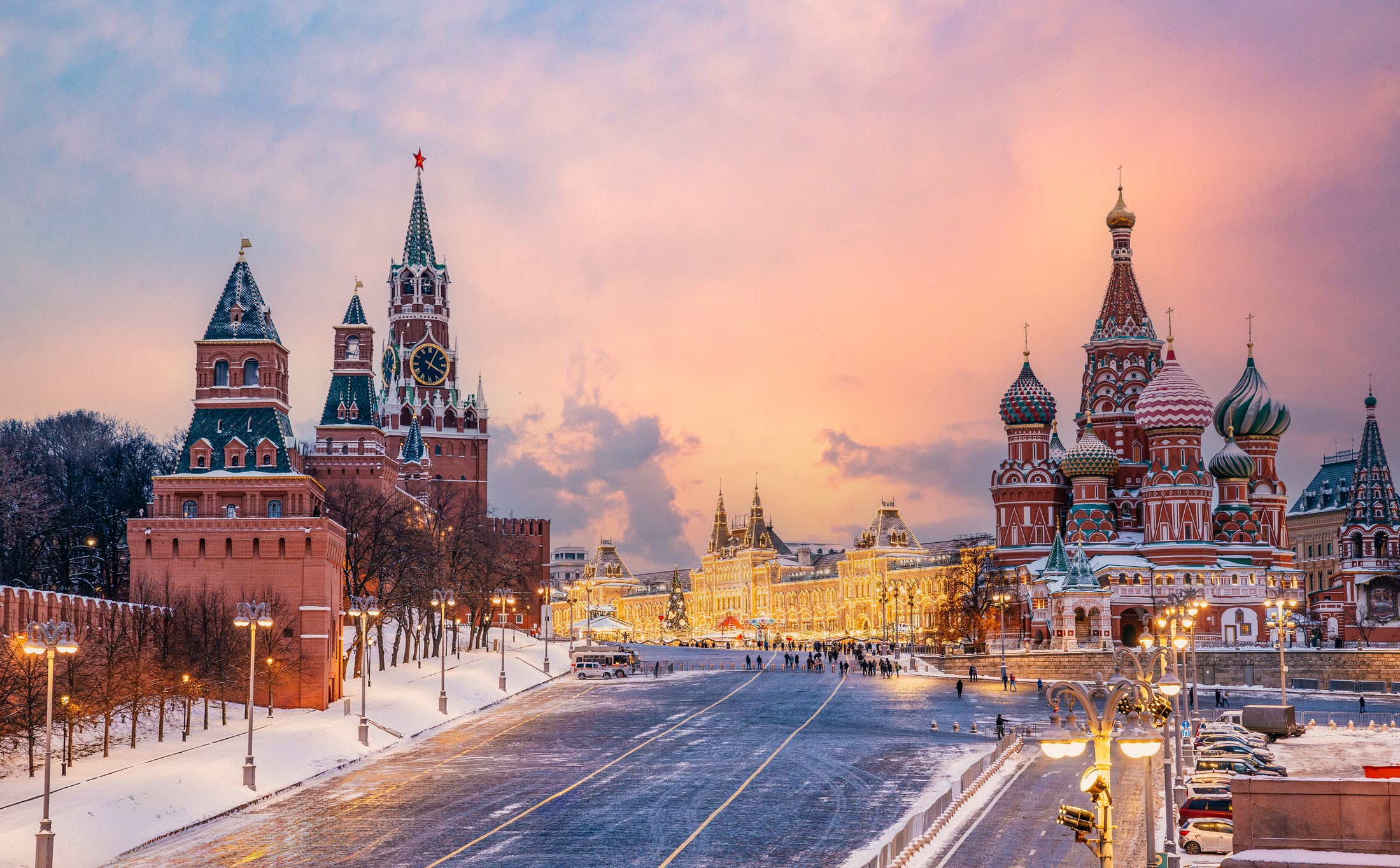 The Kremlin wall and Saint Basil's Cathedral in warm sunset hues, surrounded by a snow-covered Red Square with soft lighting and faint orange skies in the background.