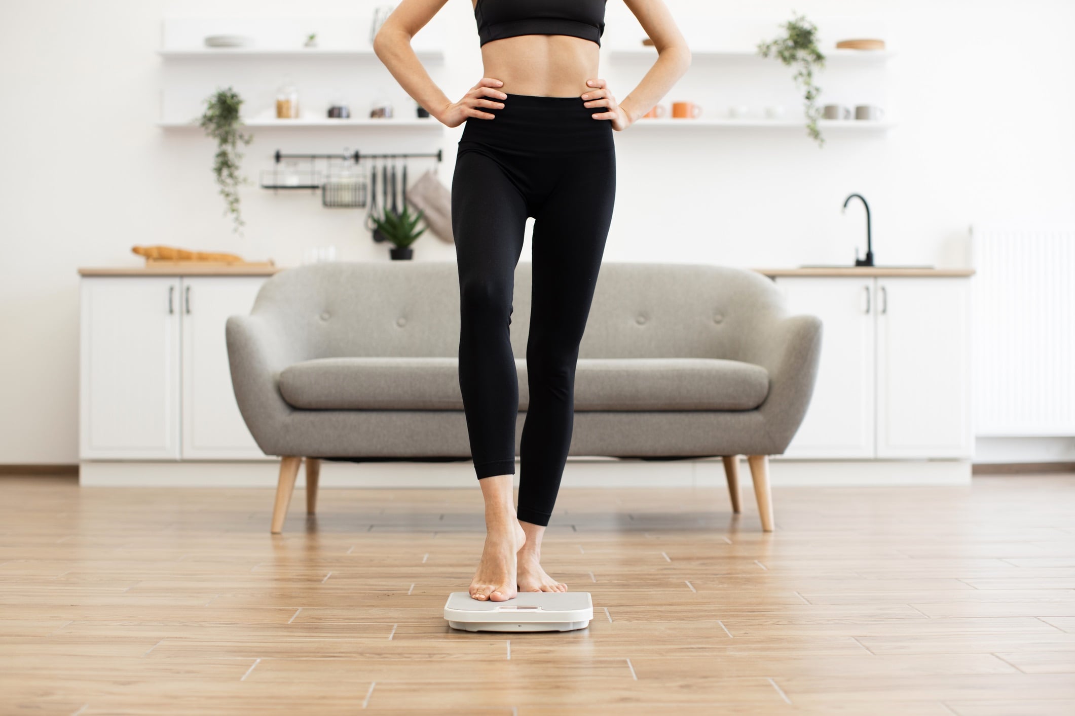 Fit woman wearing workout clothes stepping on digital scale in bright modern living room. Concept of healthy lifestyle, weight management, fitness goals, and home exercise.