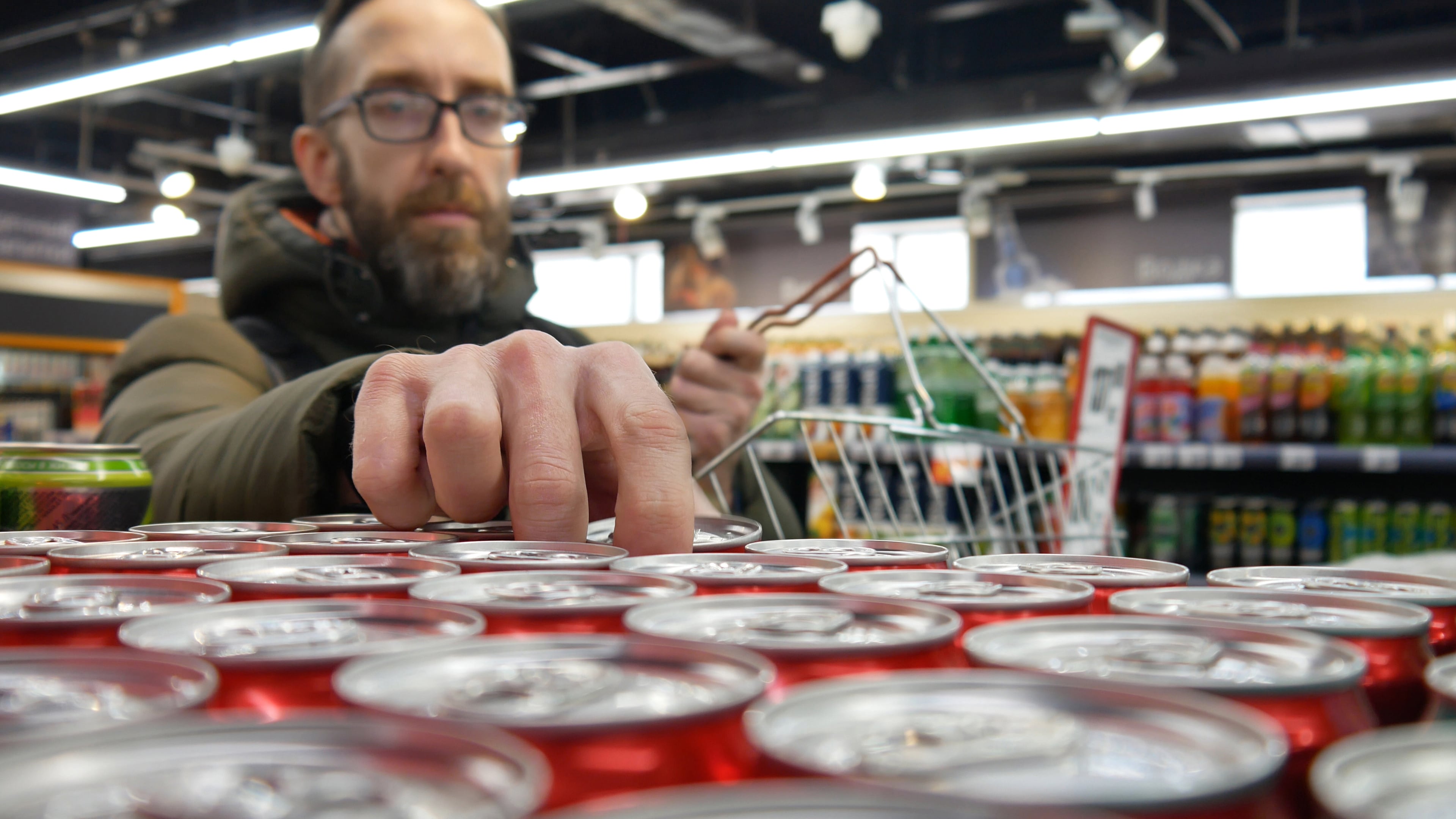 A male buyer with a shopping basket takes a few red cola cans on a supermarket shelf