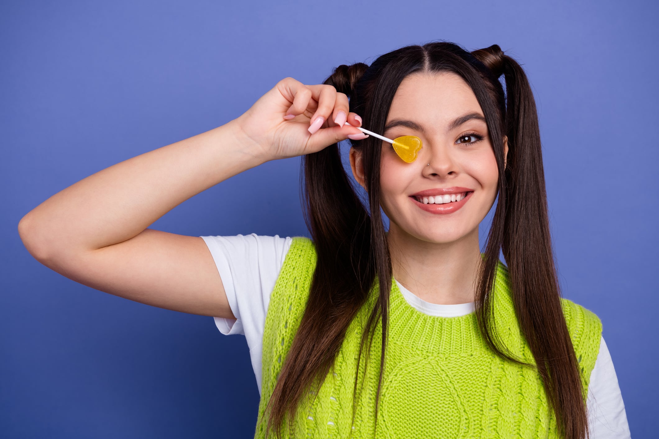 Young woman wearing lime green knit vest and white shirt, and holding a lollipop over one eye. Purple background.