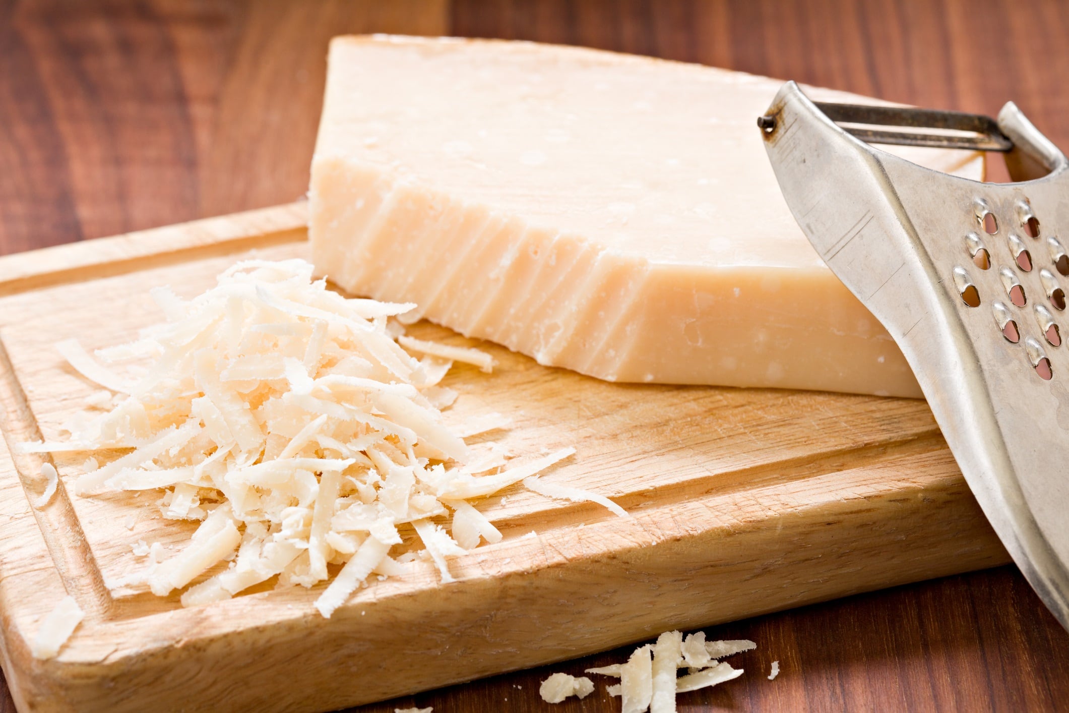 A high angle close up of a partially grated piece of Parmesan cheese on a wooden cutting board, and a small metal grater.