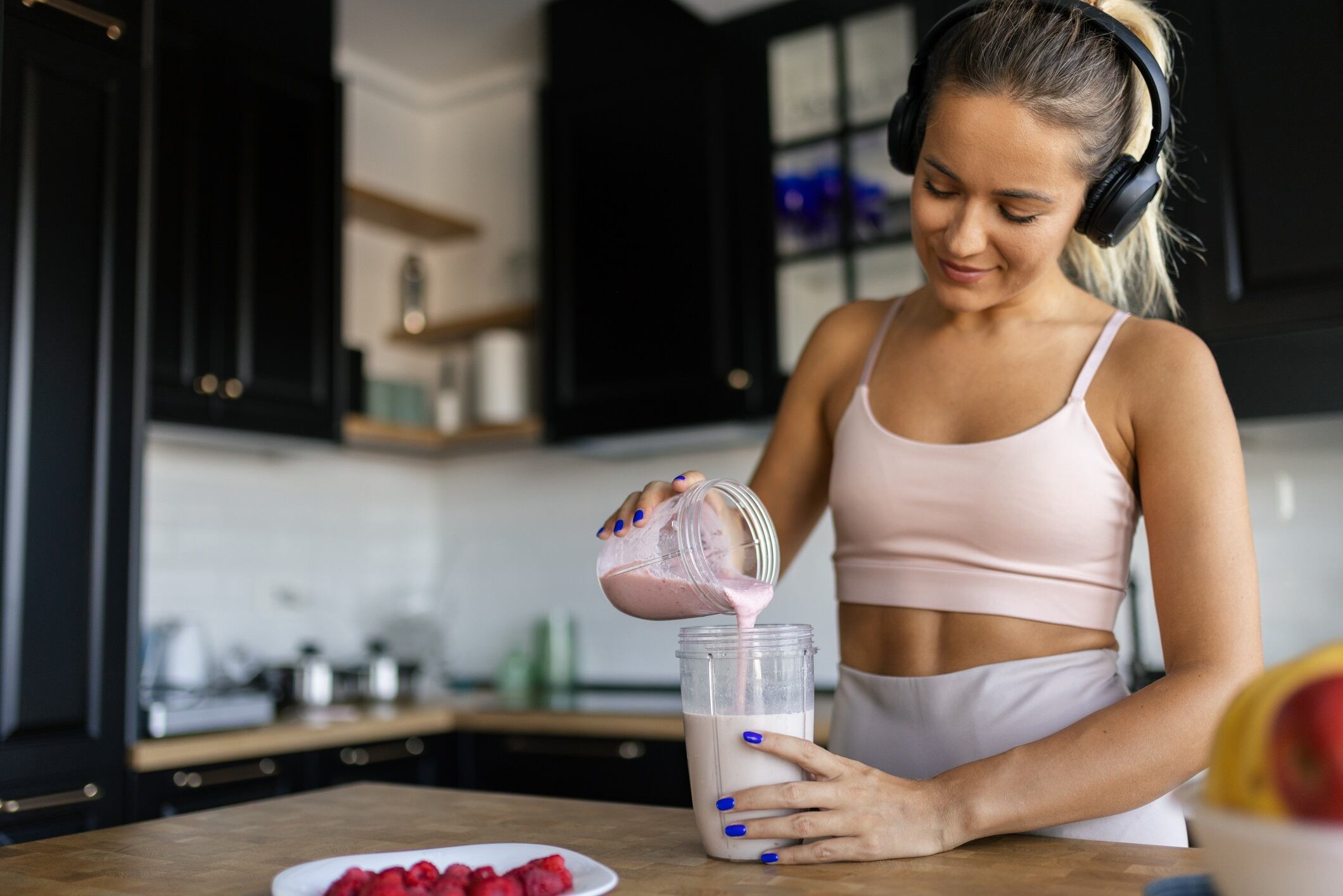 Attractive mid-adult woman dressed in sports clothes, preparing smoothie in her kitchen, getting ready for a new day. She is wearing headphones, listening to music