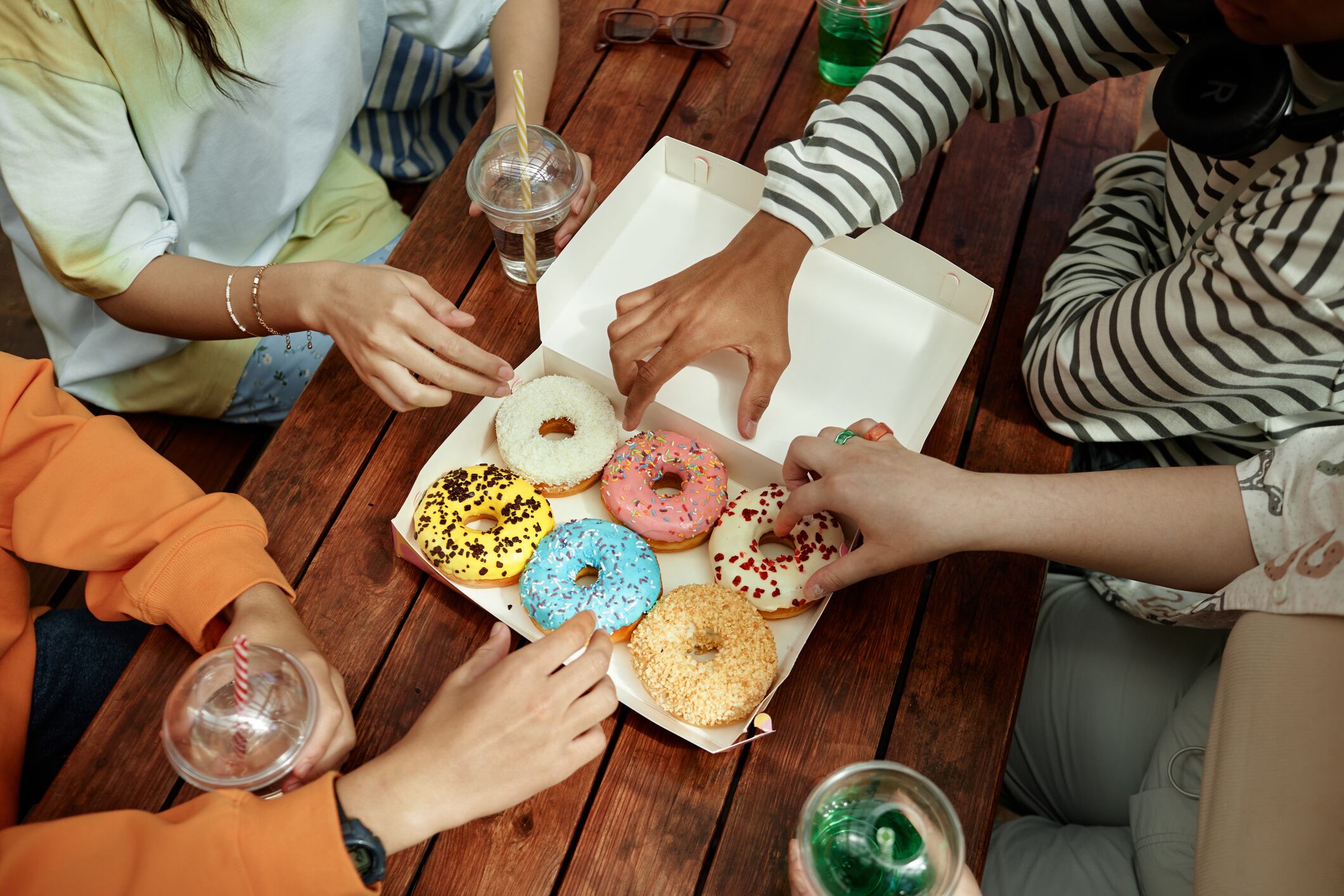 High angle shot of unrecognizable teenagers sitting at table in park cafe reaching for one of delicious glazed donuts arranged in takeaway packaging