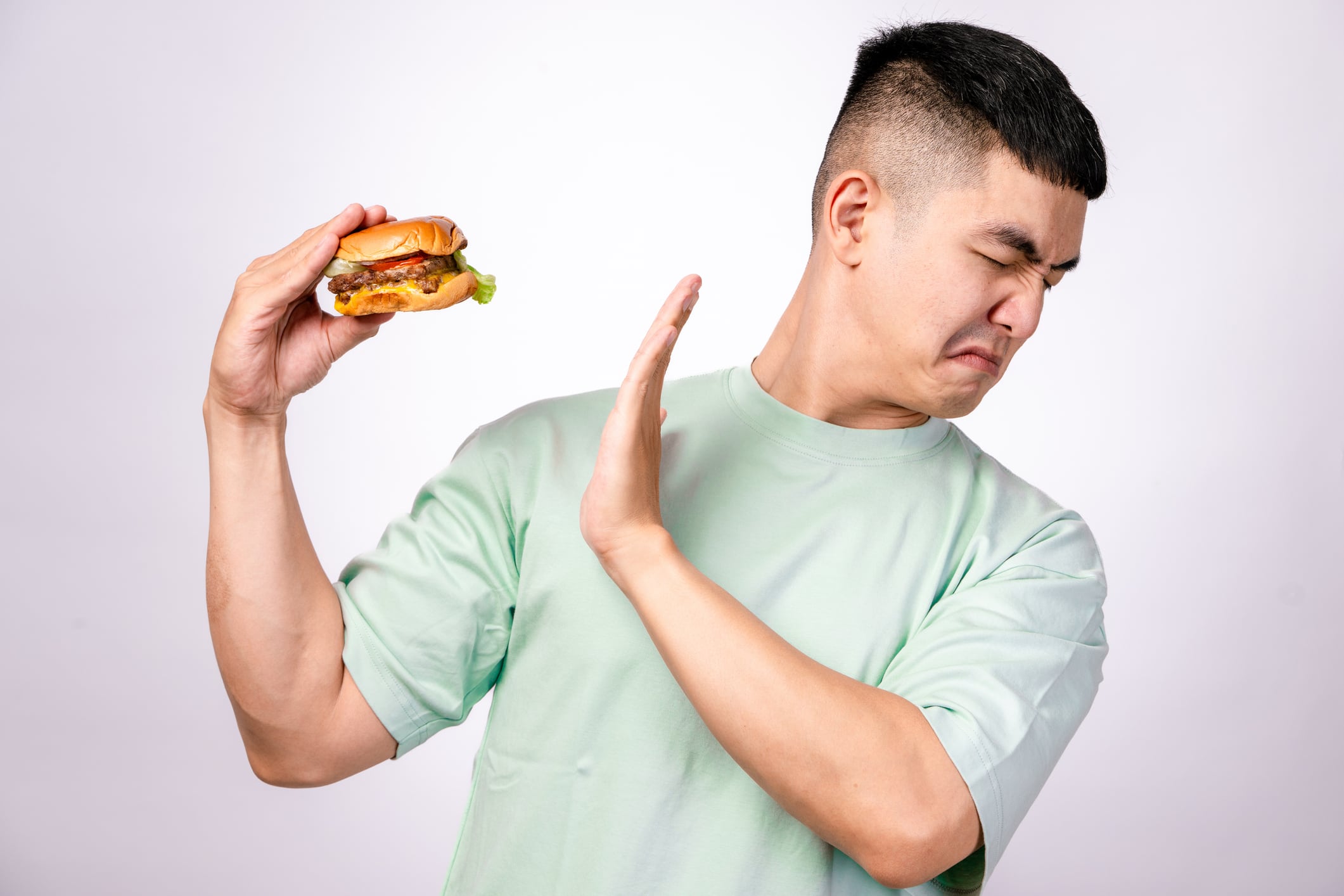 An Asian man in a mint green shirt holds a plate with a cheeseburger while extending his other hand in a stop gesture. He looks surprised or disapproving. The background is plain white