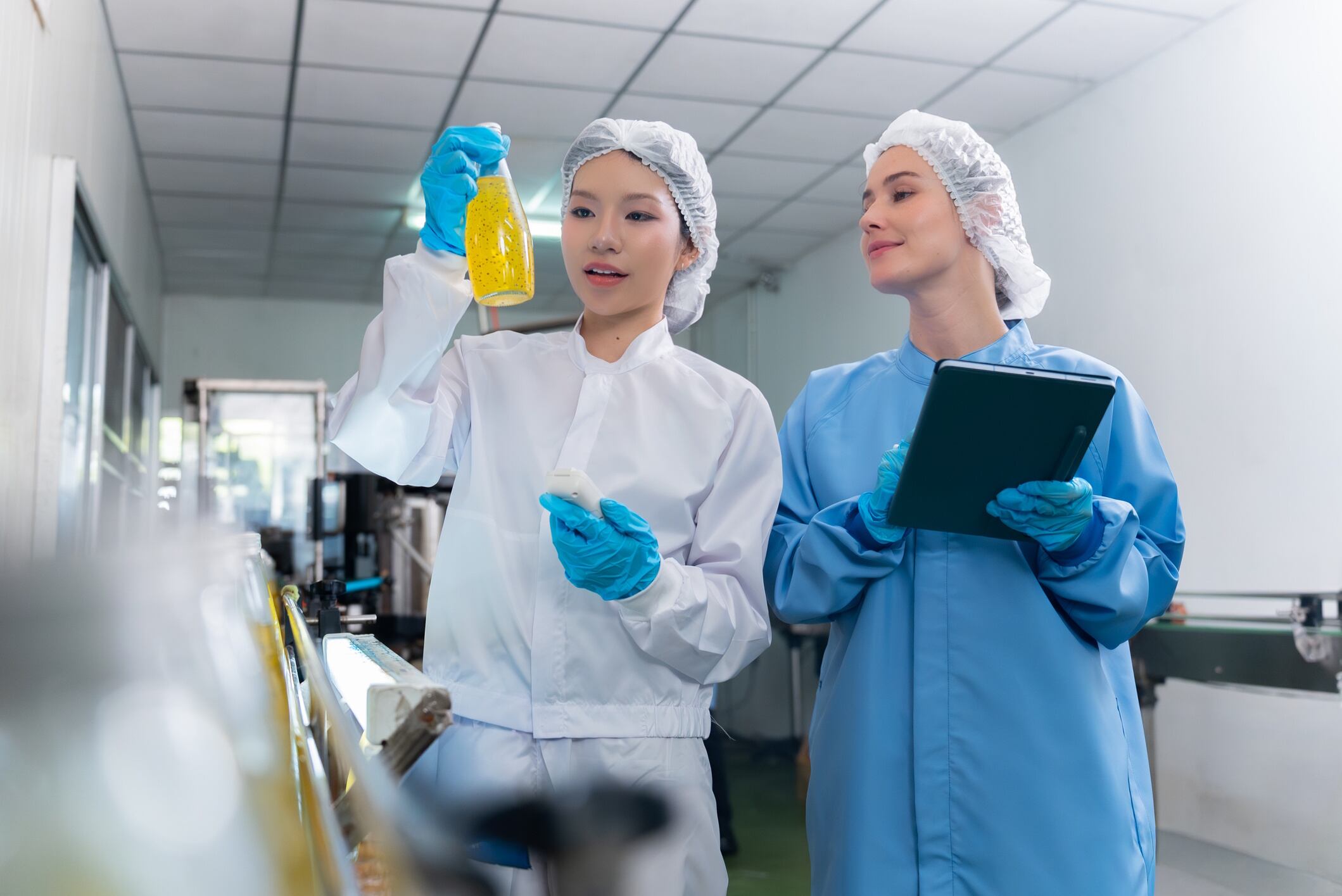 Two young adult women scientists in protective clothing inspect a yellow liquid sample in a laboratory or food processing facility mith_67 GettyImages