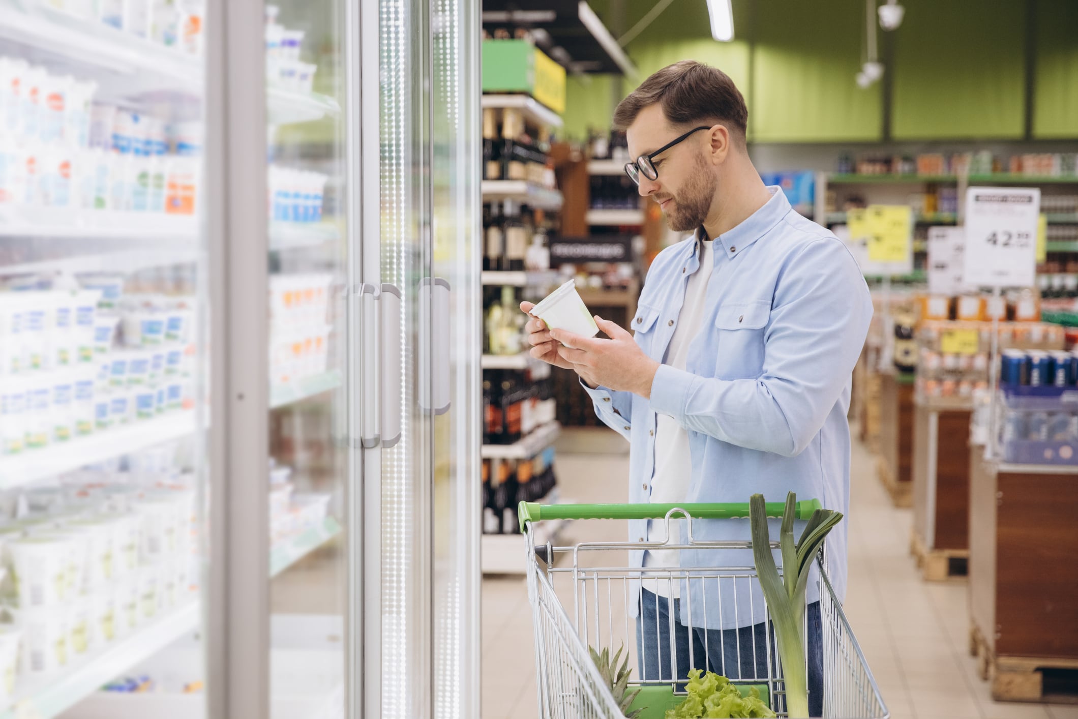 Man choosing groceries, reading product information on a package in front of refrigerated section in a supermarket