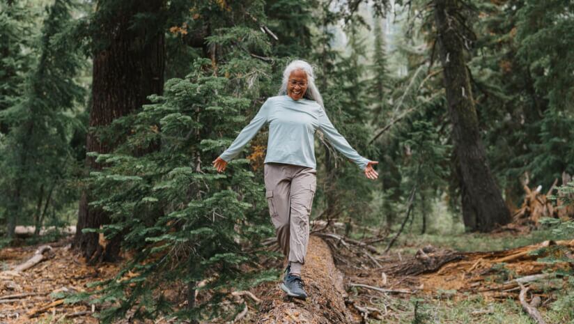 An older woman walking in the forest balancing on a fallen tree trunk