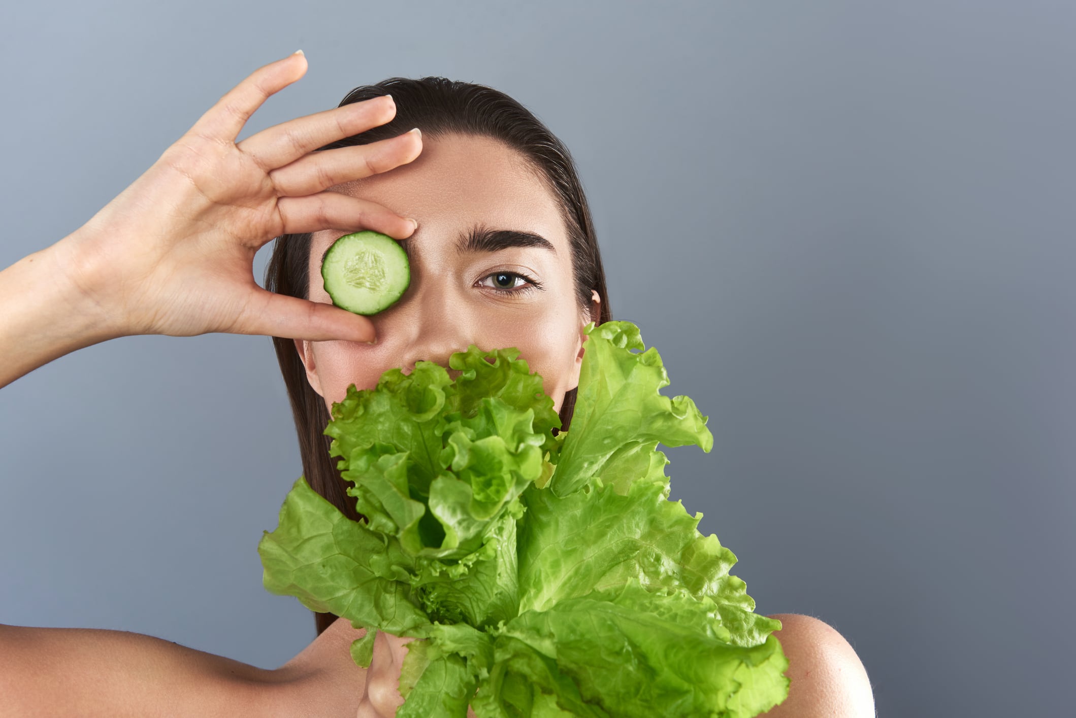Portrait of attractive lady standing and looking at camera. She is hiding mouth with salad and eye with cucumber