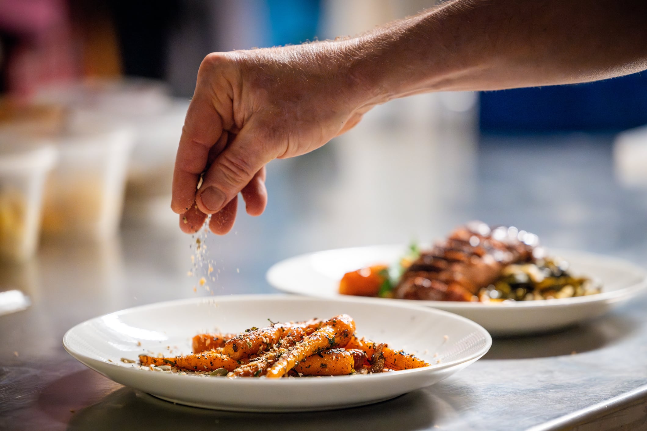 A hand stretches into the middle of frame and puts the final spices onto a dish of gleaming carrots. The plate sits on a commercial kitchen countertop. The plate of carrots and hand are in focus in the foreground, in the background sits another gourmet dish slightly out of focus.