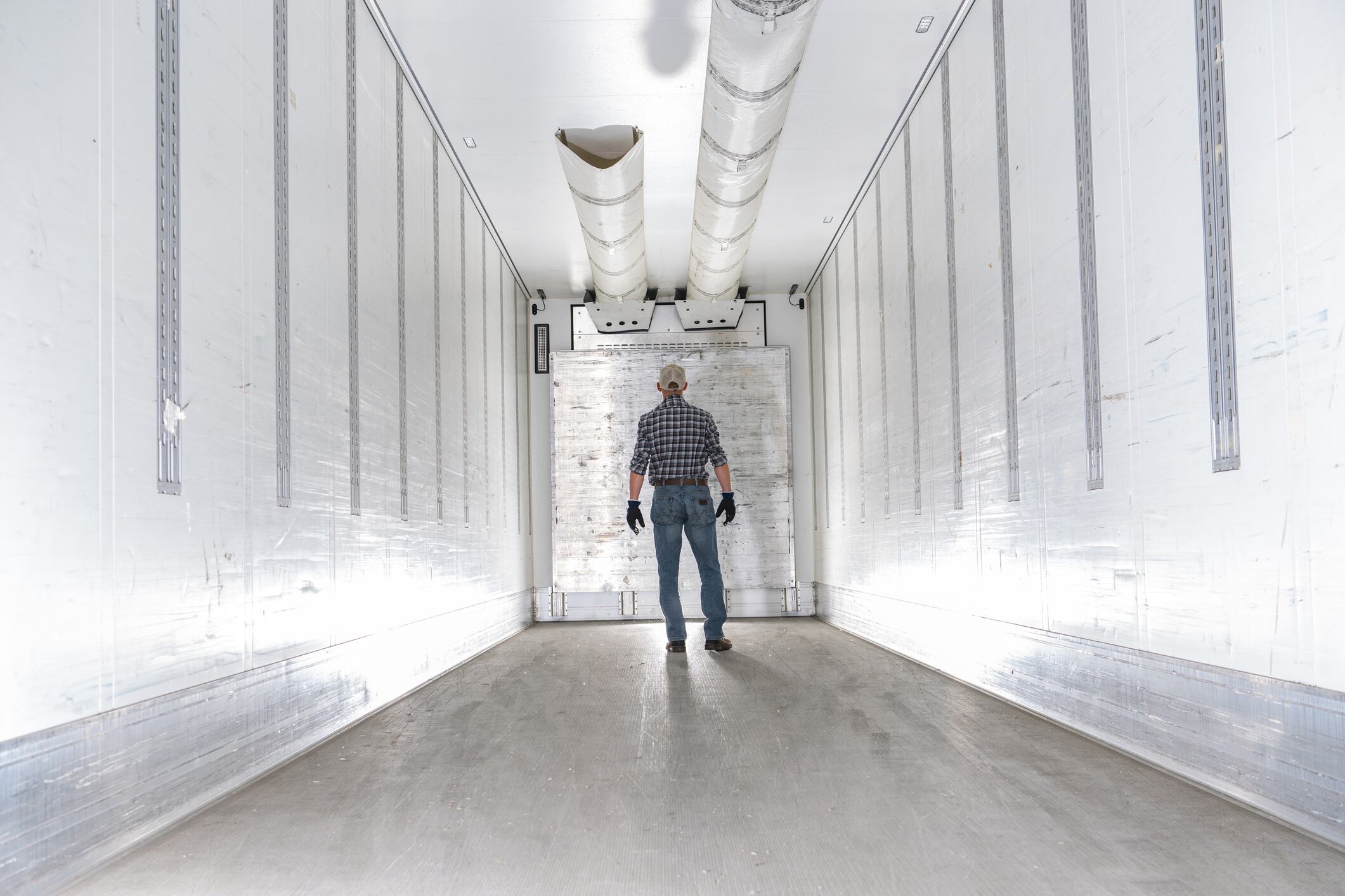 A worker in a checkered shirt and gloves stands in the middle of a refrigerated truck trailer. The interior is white and spacious, with visible cooling equipment on the ceiling. The worker appears focused as he inspects the area, ensuring it is clean and ready for cargo transport.