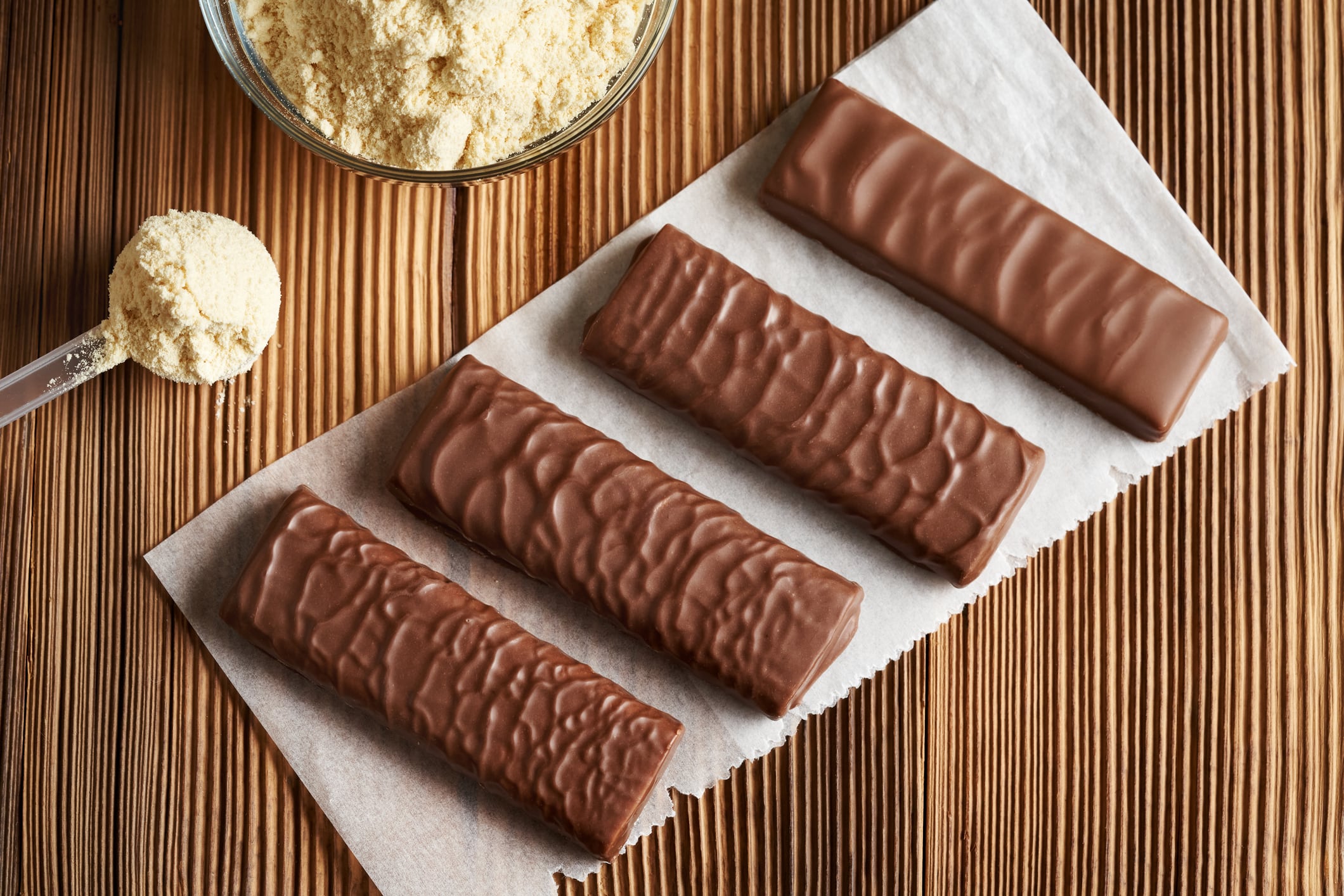 Four vegetarian protein chocolate bars with a bowl and scoop of whey protein powder on a wooden table