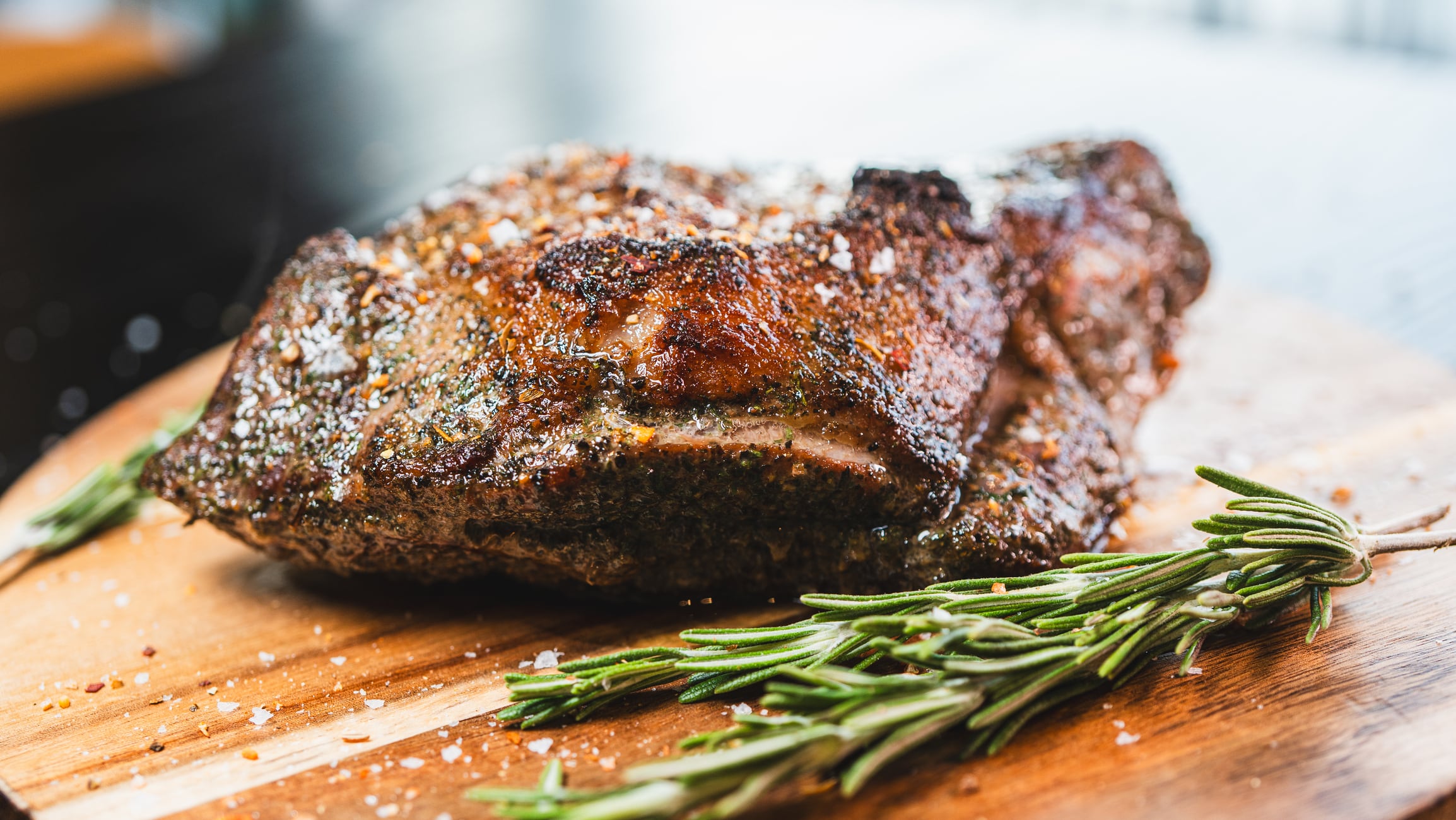 A beautifully presented, whole roasted leg of lamb, perfectly cooked and resting on a wooden board. Garnished with fresh rosemary and herbs, this succulent centerpiece is ideal for a festive meal or celebration.