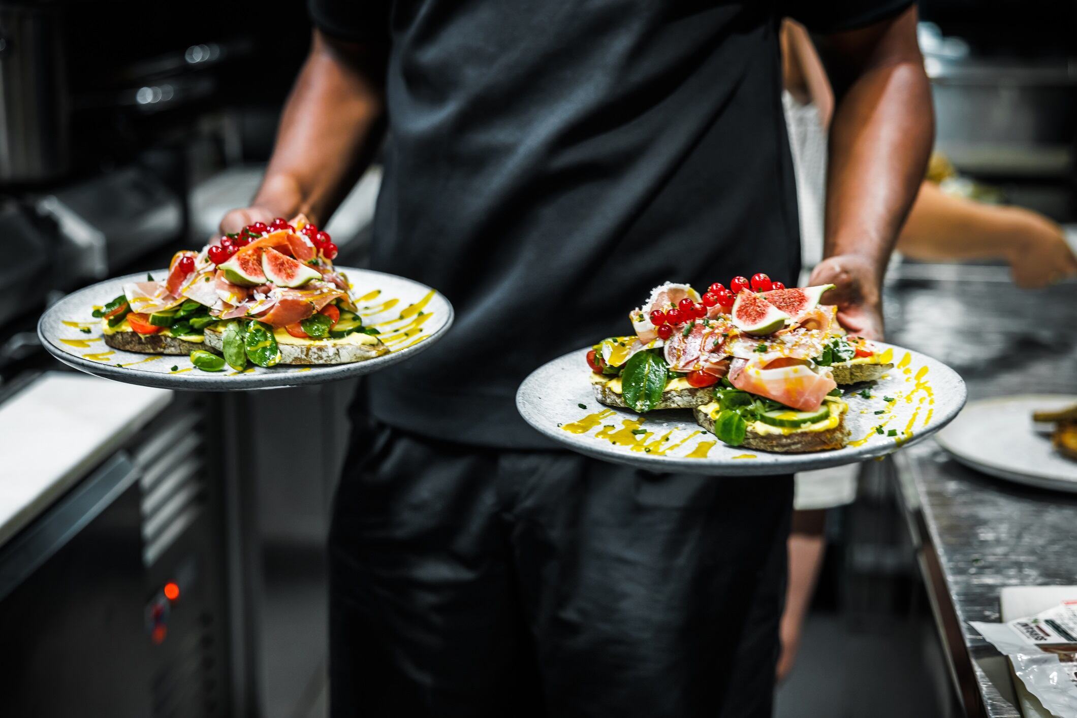 In a bustling kitchen, a chef presents two plates of beautifully garnished avocado toasts, ready to be served to awaiting customers.