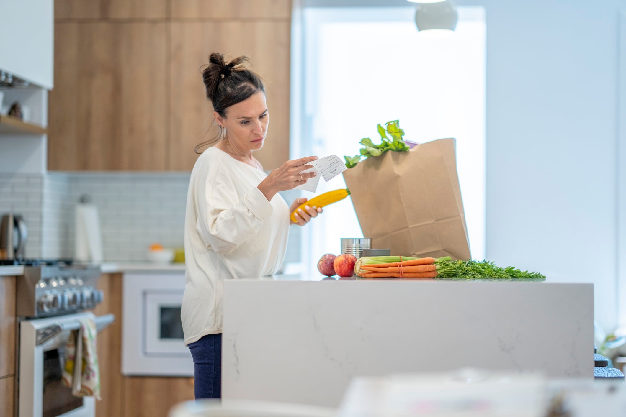 A young woman stands at the kitchen counter holding her grocery bill as she reviews it with disbelief at the rising cost of food.  Her groceries are sitting in a paper bag on the counter in front of her and she is dressed casually.