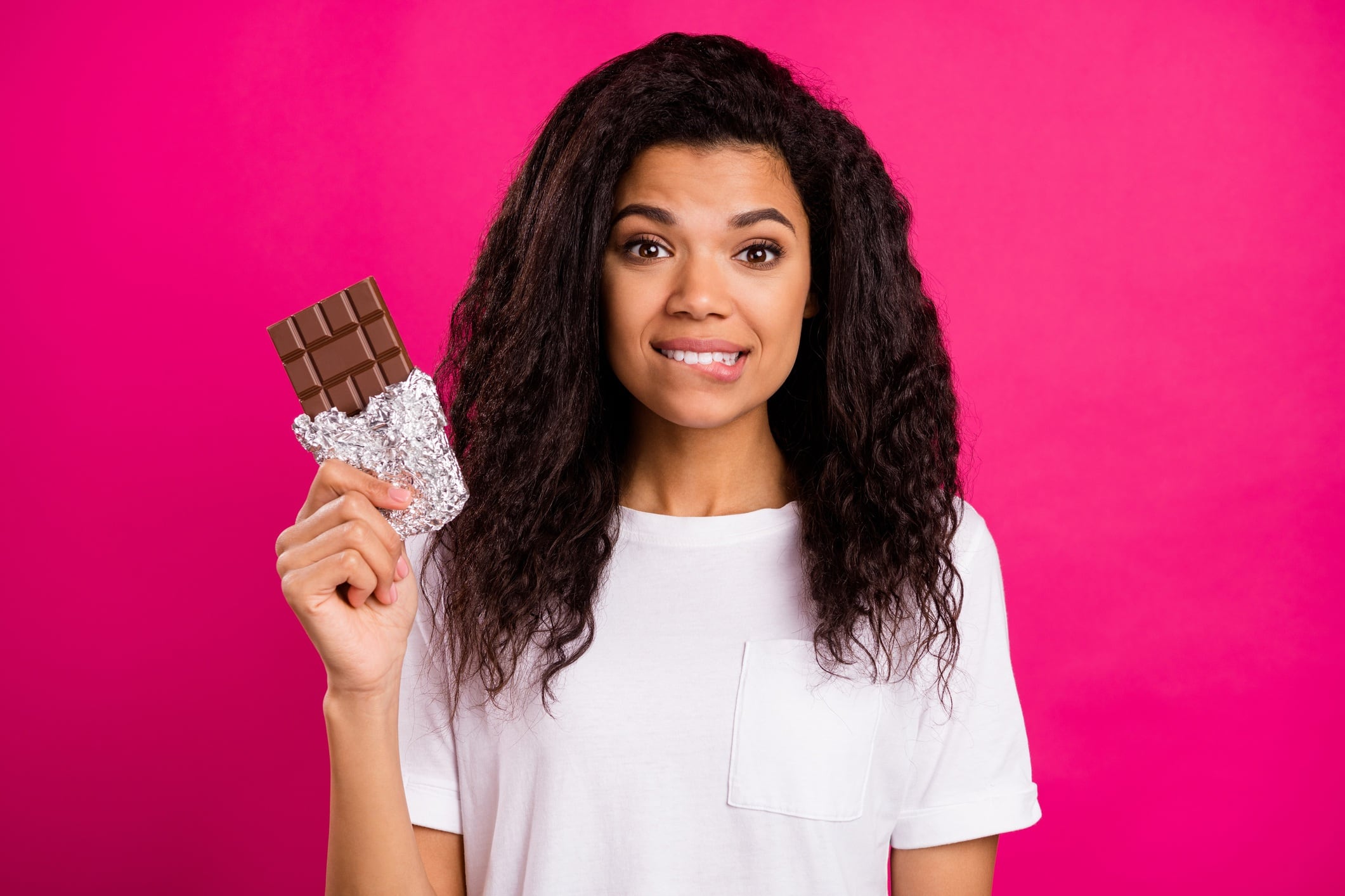 Woman holding chocolate bar. She's wearing a white t-shirt and is isolated on pink coloured background.