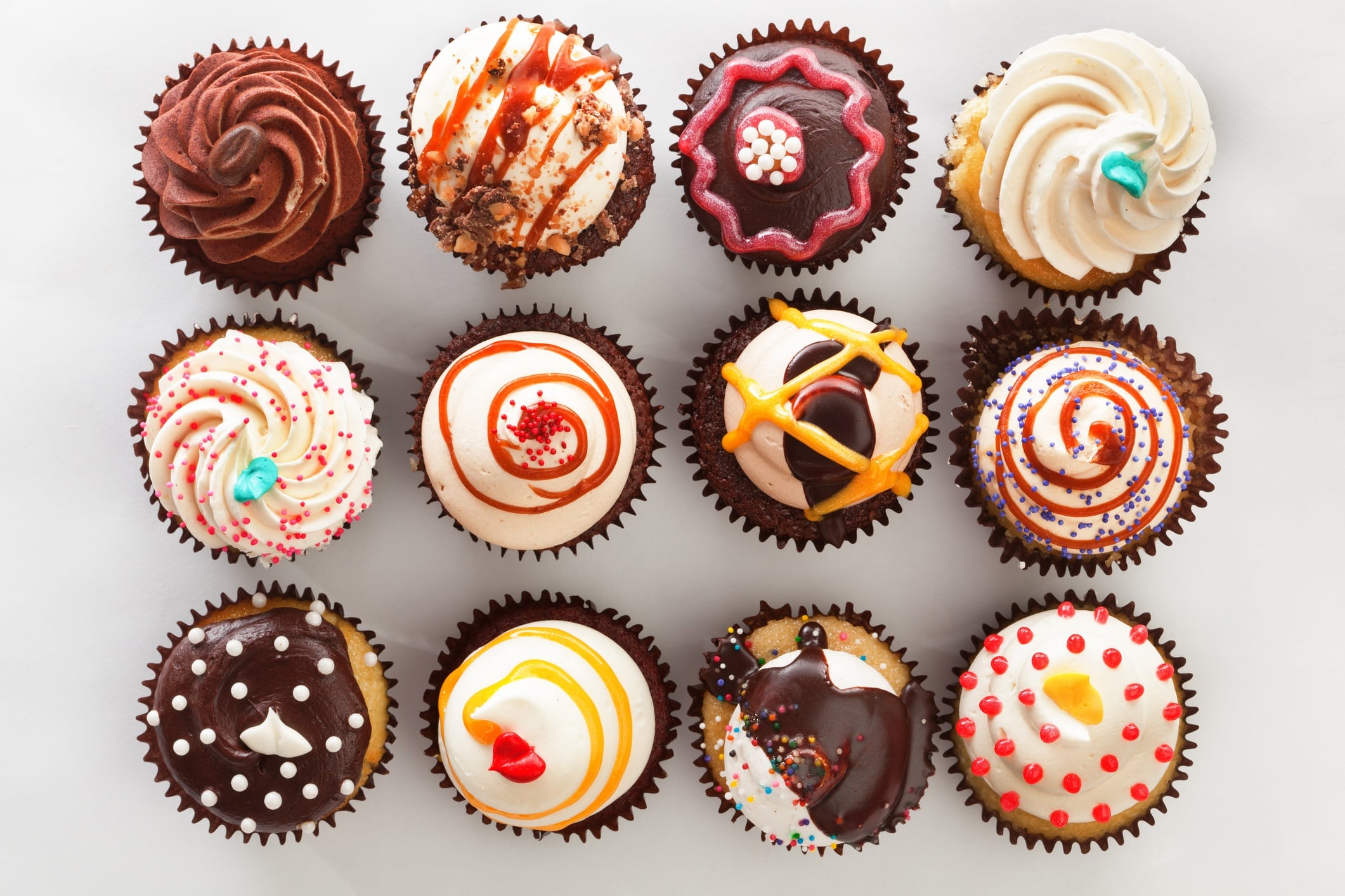 Horizontal high angle, directly overhead view of a dozen frosted cupcakes. The cupcakes are against a white background and are decorated with swirls, sprinkles, drizzles and candies—a tempting decadent treat for those who indulge in sweets and love cakes with lots of toppings.