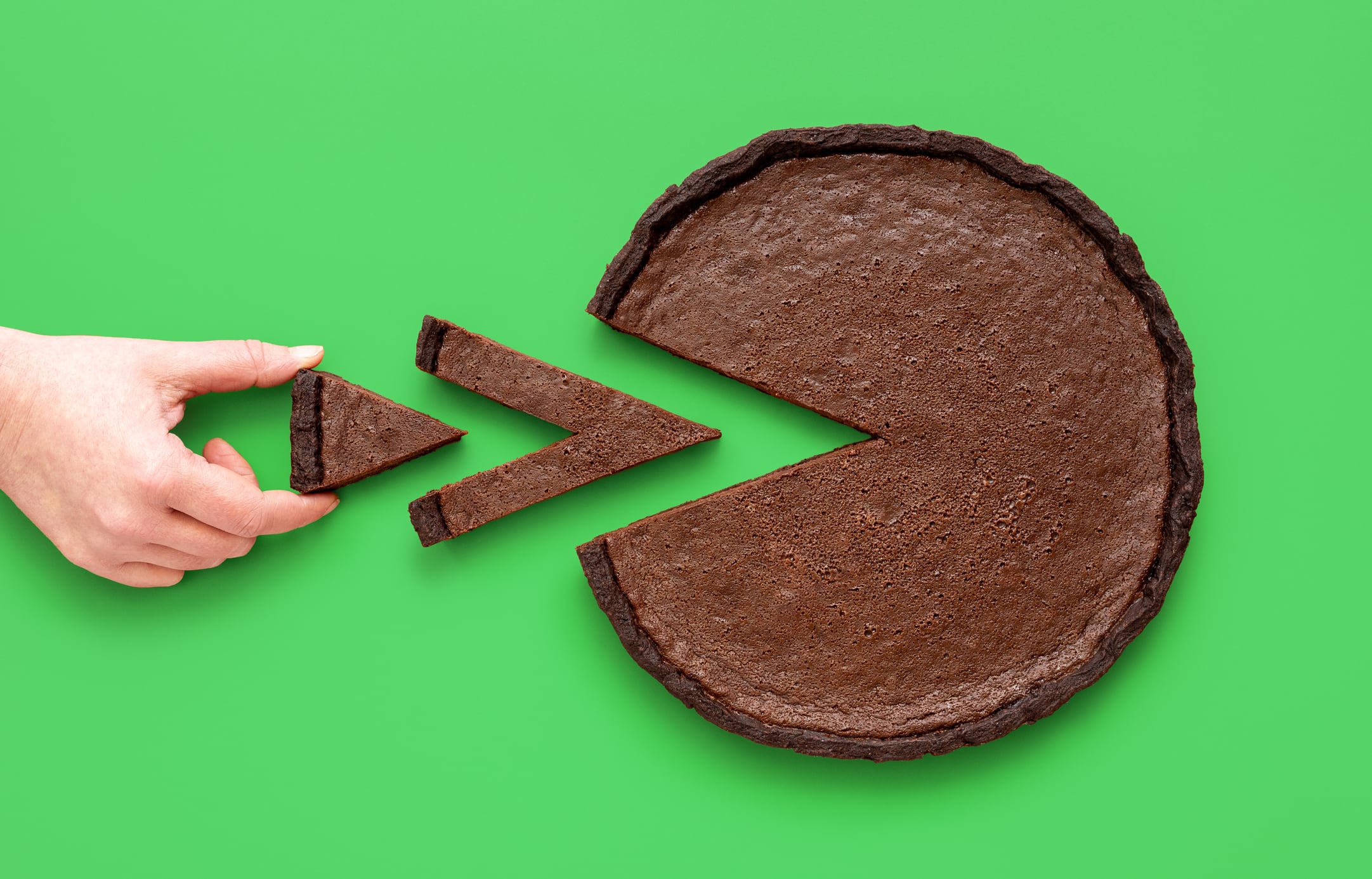 Above view with a woman's hand taking a tiny slice of chocolate tart, minimalist on a green background.