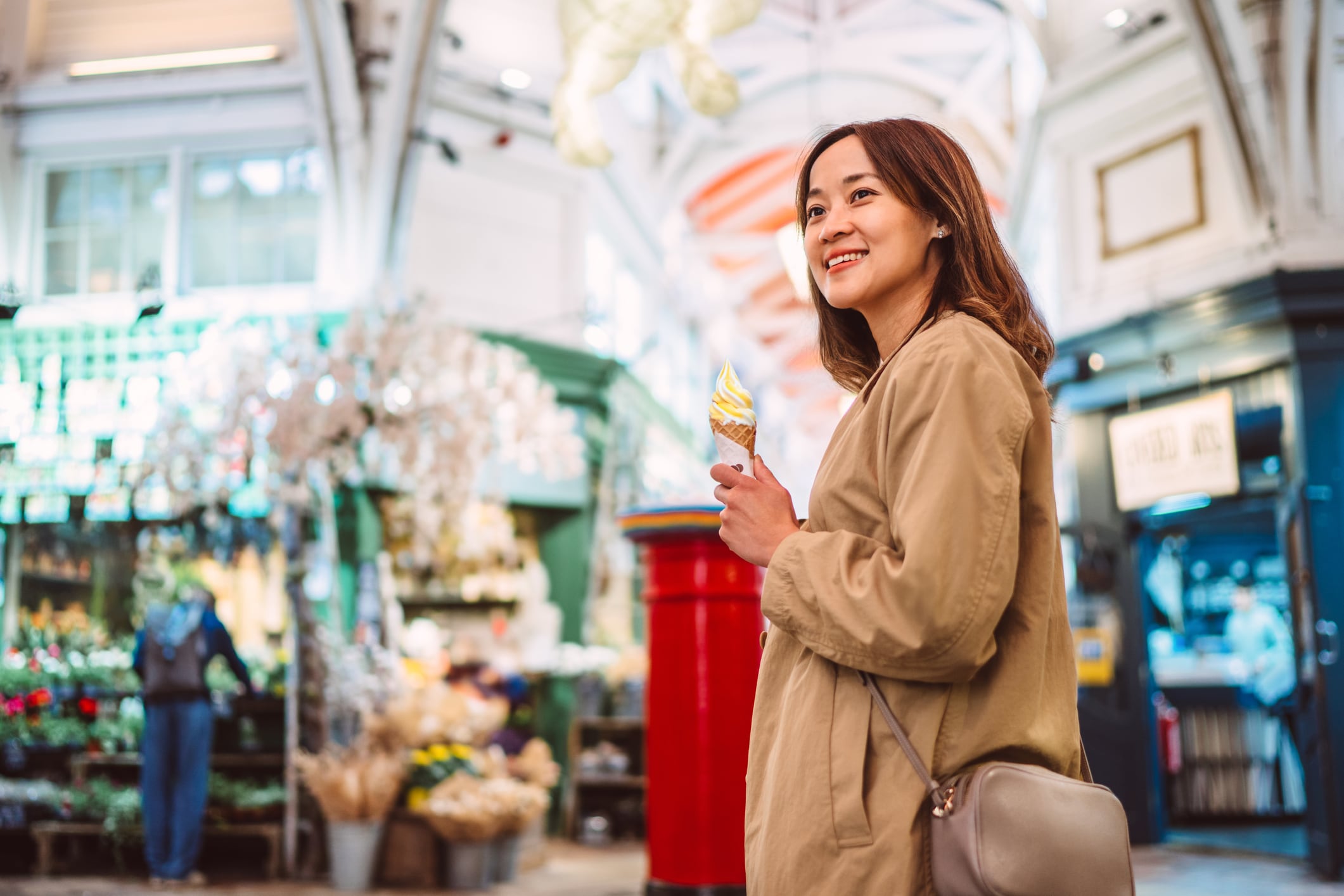 Cheerful woman holds a swirl ice cream cone while walking through a bustling indoor market filled with shops and decorations. She looks happy and relaxed, enjoying a leisure moment. Perfect for concepts of lifestyle, city exploration, local culture, weekend activities, urban tourism, and enjoying simple pleasures.