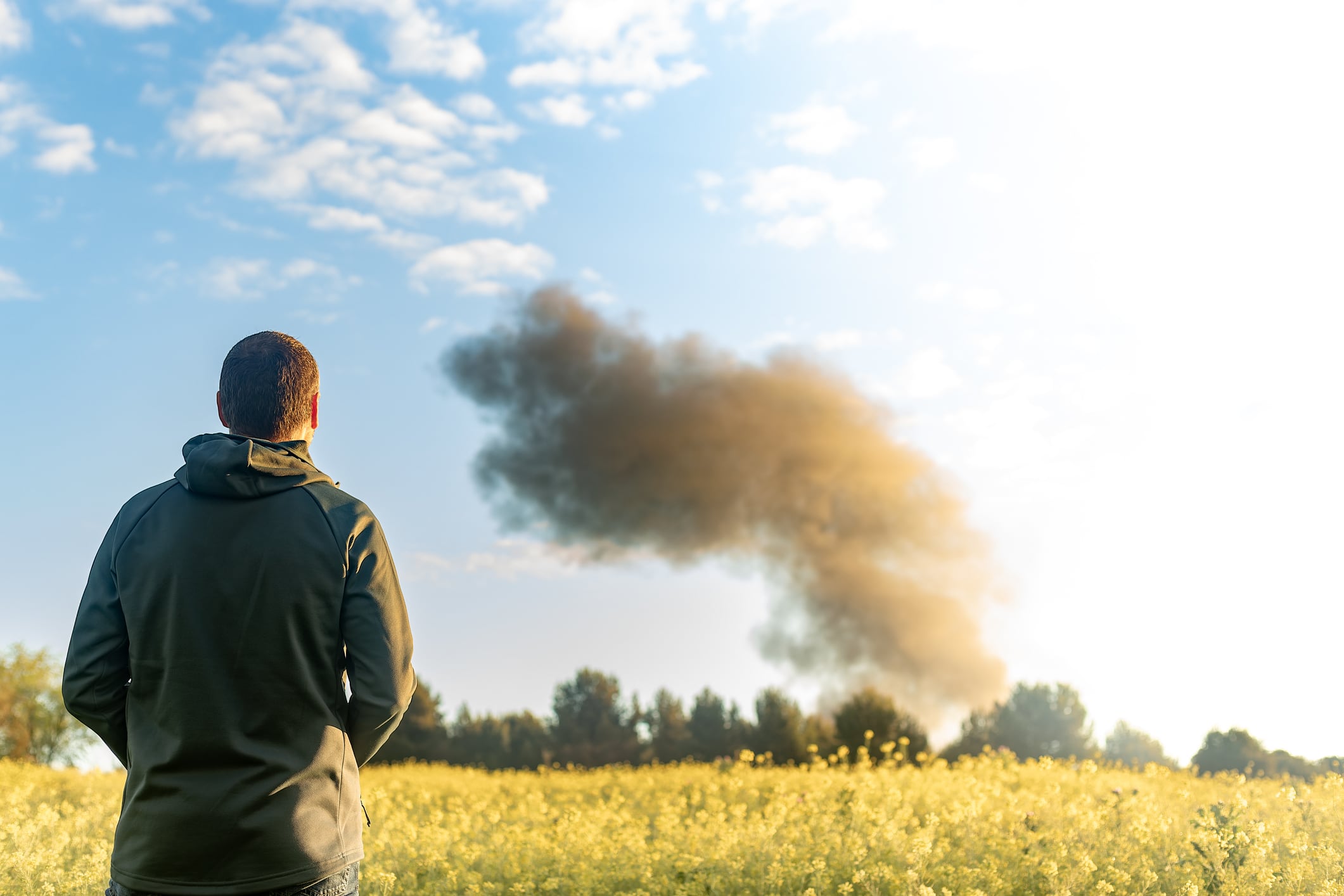 Alcorcón. Madrid. Spain. Rear view of a farmer standing in a rapeseed field, watching a large column of black smoke rising from a distant fire, causing air pollution and environmental damage.