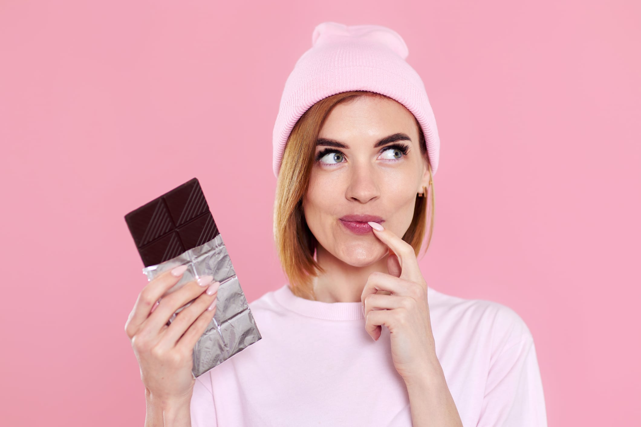 Blonde woman in pink t-shirt and hat, holding chocolate bar, on pink background.