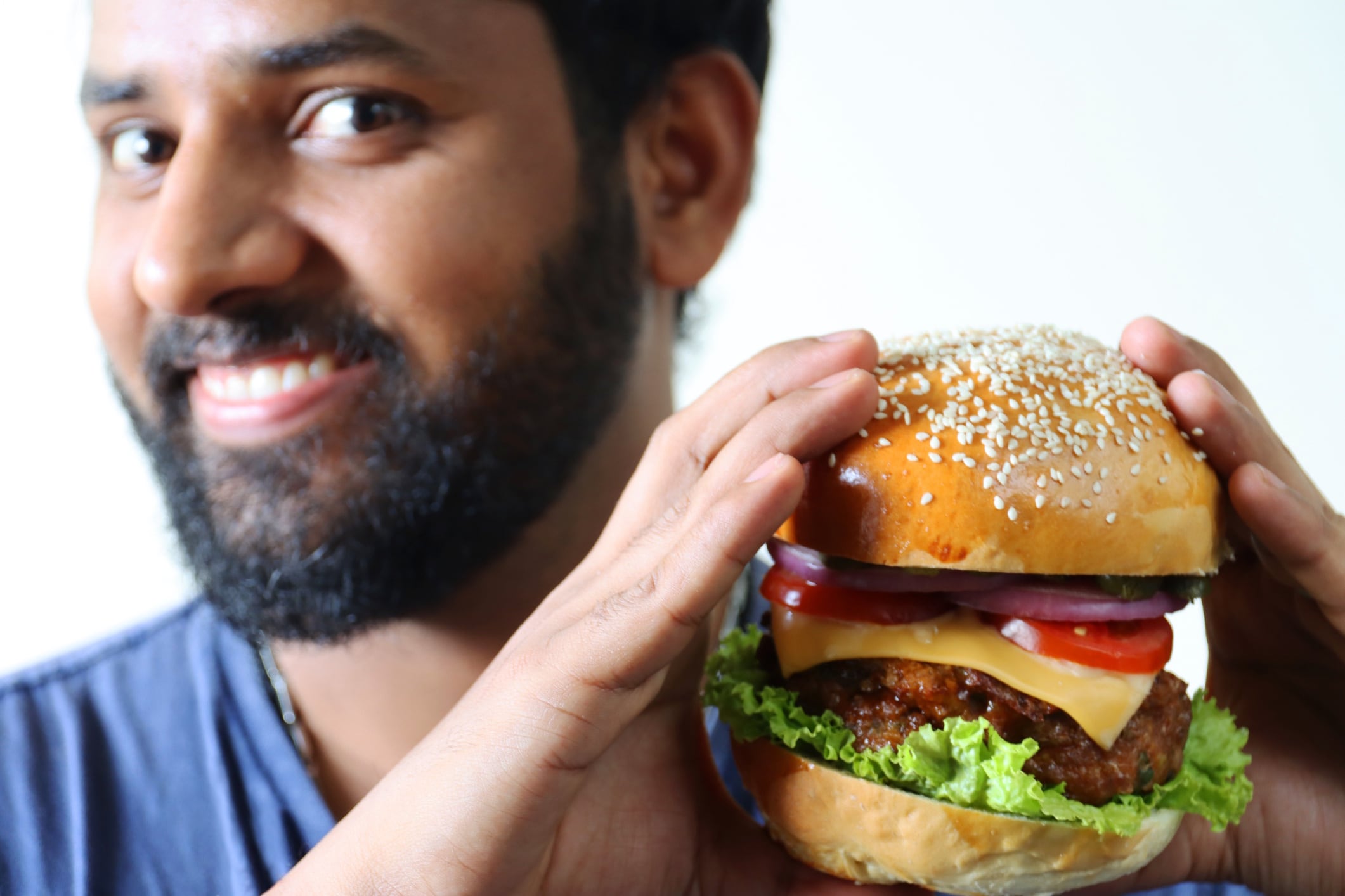 Image of smiling, asian man holding gourmet, homemade cheese burger, bread bun with sesame seeds, melted cheese, sliced tomato, gherkins, red onion rings and lettuce