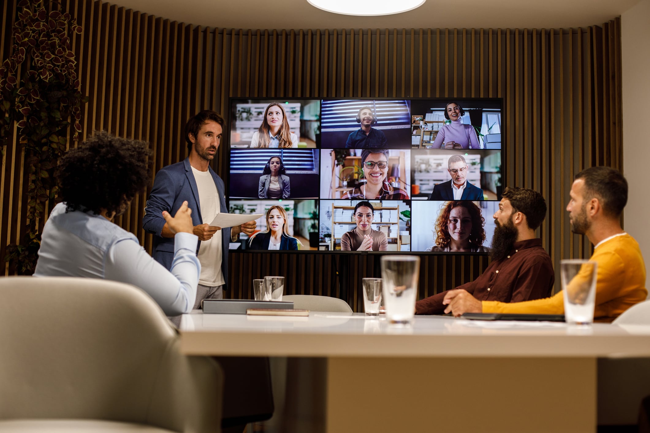 Candid shot of four businessmen sitting around conference table in a board room, having a meeting, with some colleagues on a video call, discussing project ideas and strategies.