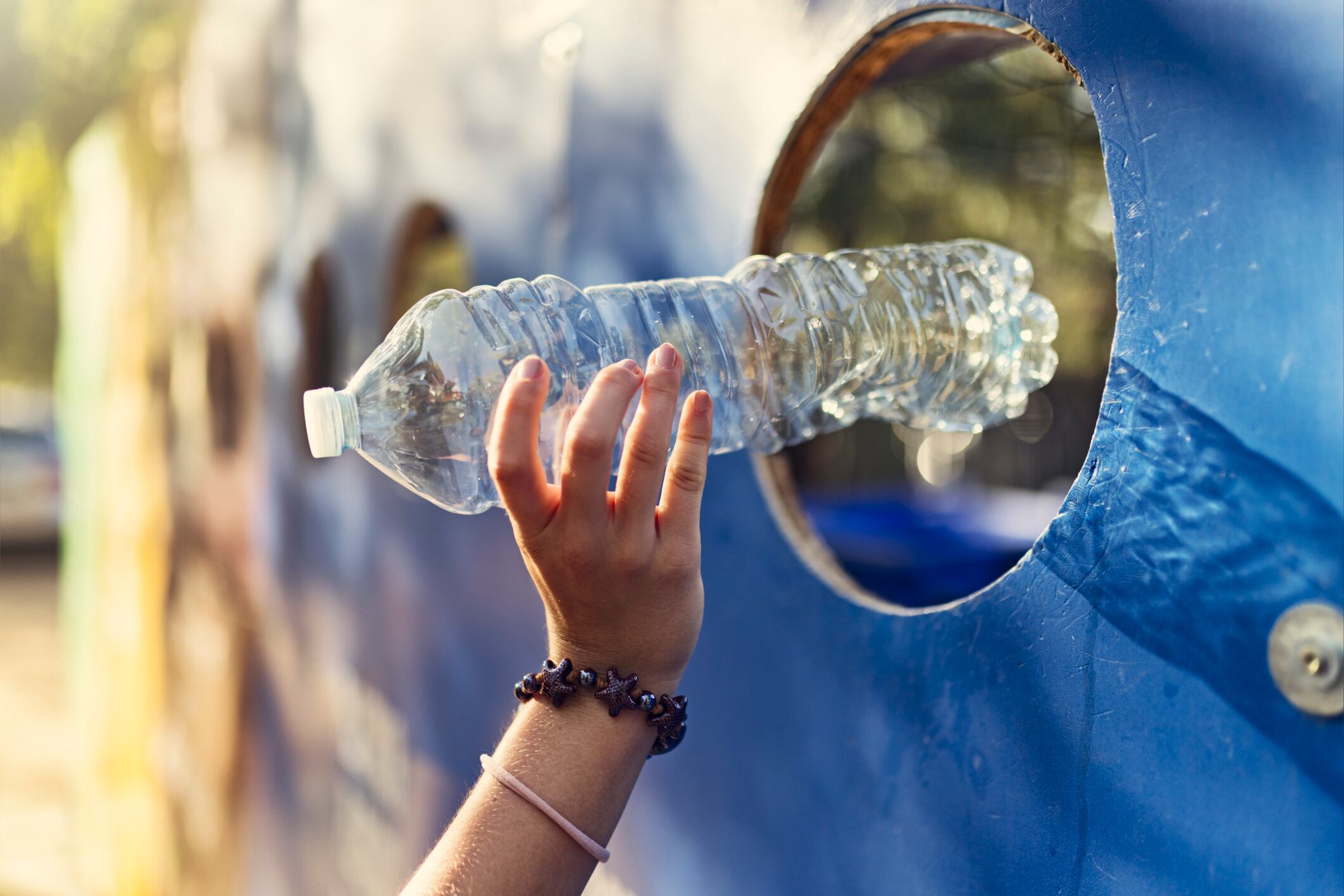 Close-up of teenage girl hand engage in responsible waste disposal at a public plastic recycling station. Teenage girl, dressed in a red shirt, is placing plastic bottles into a circular slot on a bold blue recycling wall marked with “PLÁSTICA.” 
Shot with Canon R5