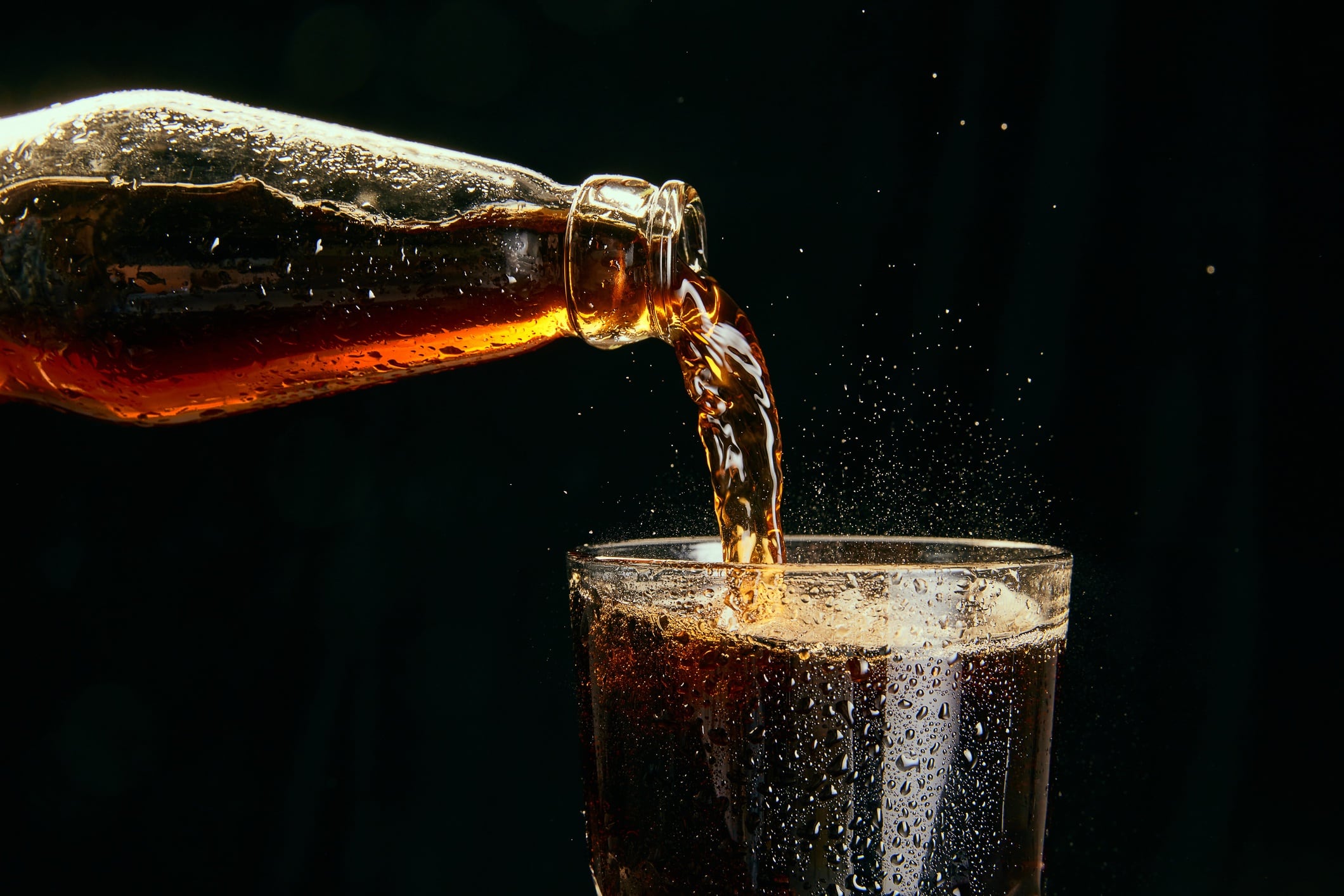 Cola pouring from bottle into glass against black background.