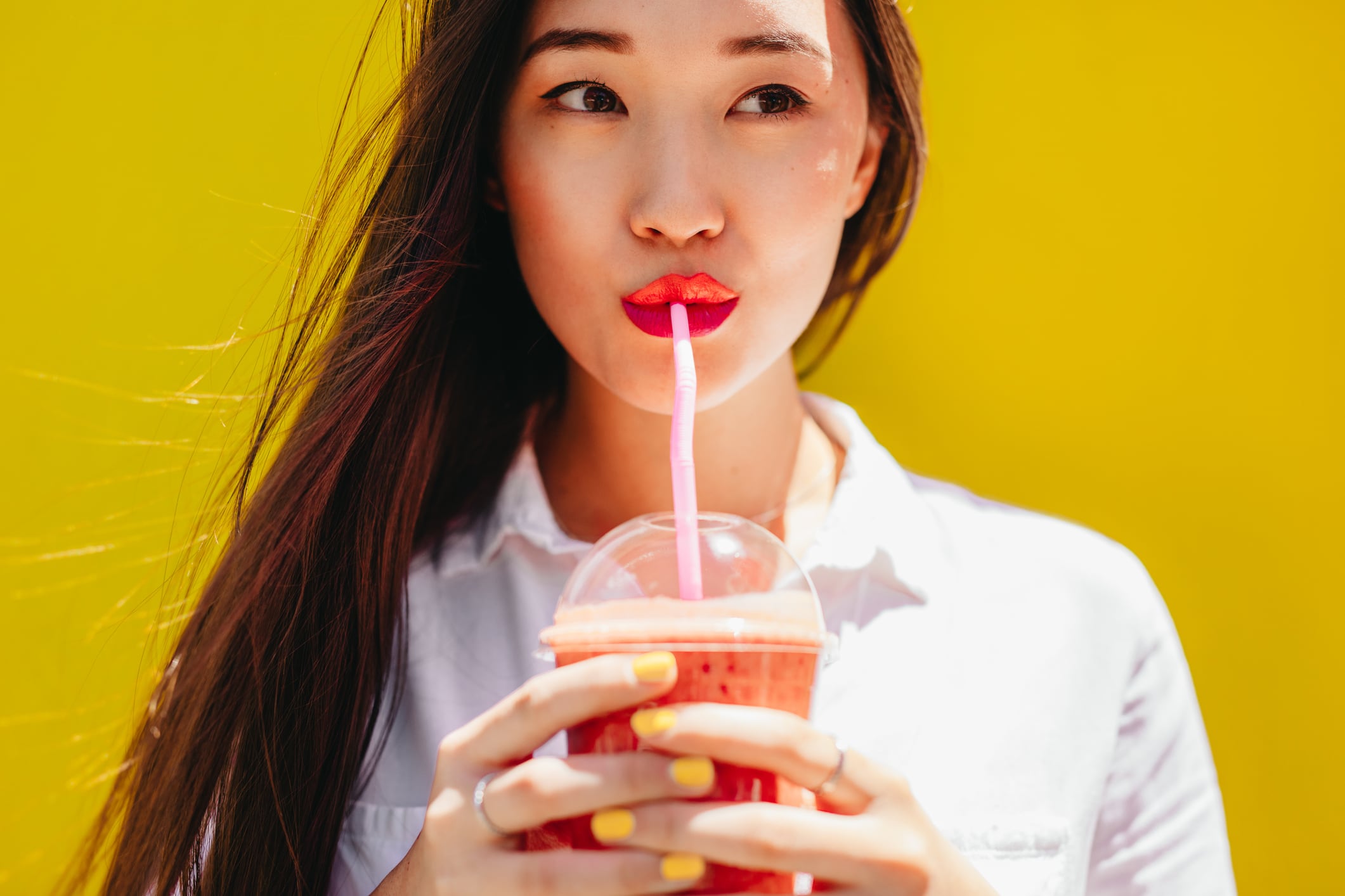 Woman drinking juice from a plastic cup using a straw. Yellow background.