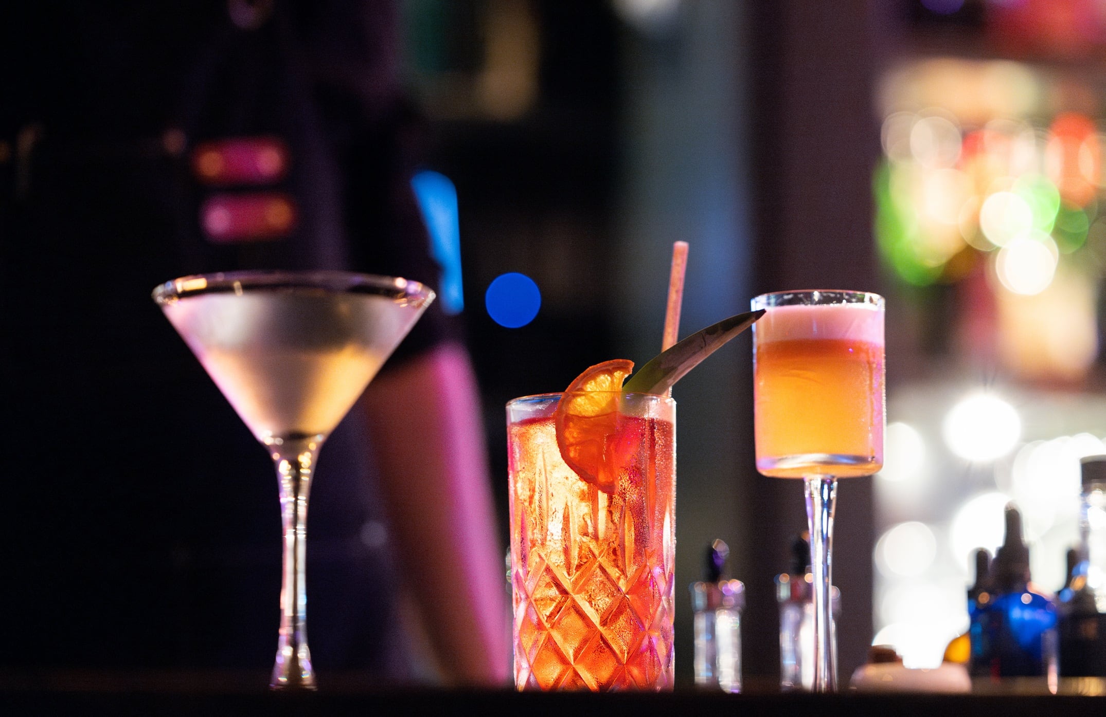 Three cocktails sit on a bar counter in a darkly lit bar. The cocktails are illuminated, a martini glass slightly out of focus on the left, and two other drinks in focus to the right. In the background is the bartender's arm, and the bright lights of the bar shelves.