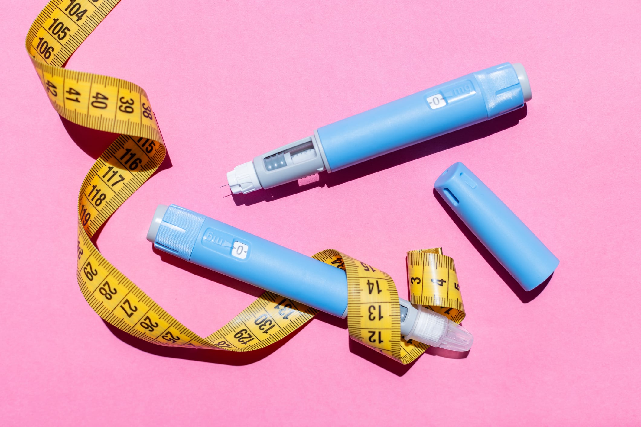 Blue Ozempic pens with yellow measuring tape on pink background. Concept of diabetes treatment, weight loss aid, and modern healthcare solutions.Yellow measuring tape around the injectors.