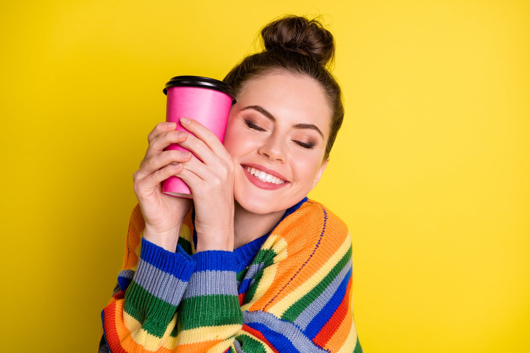 Woman wearing brightly coloured jumper and holding coffee cup close to face, while smiling. Yellow coloured background.