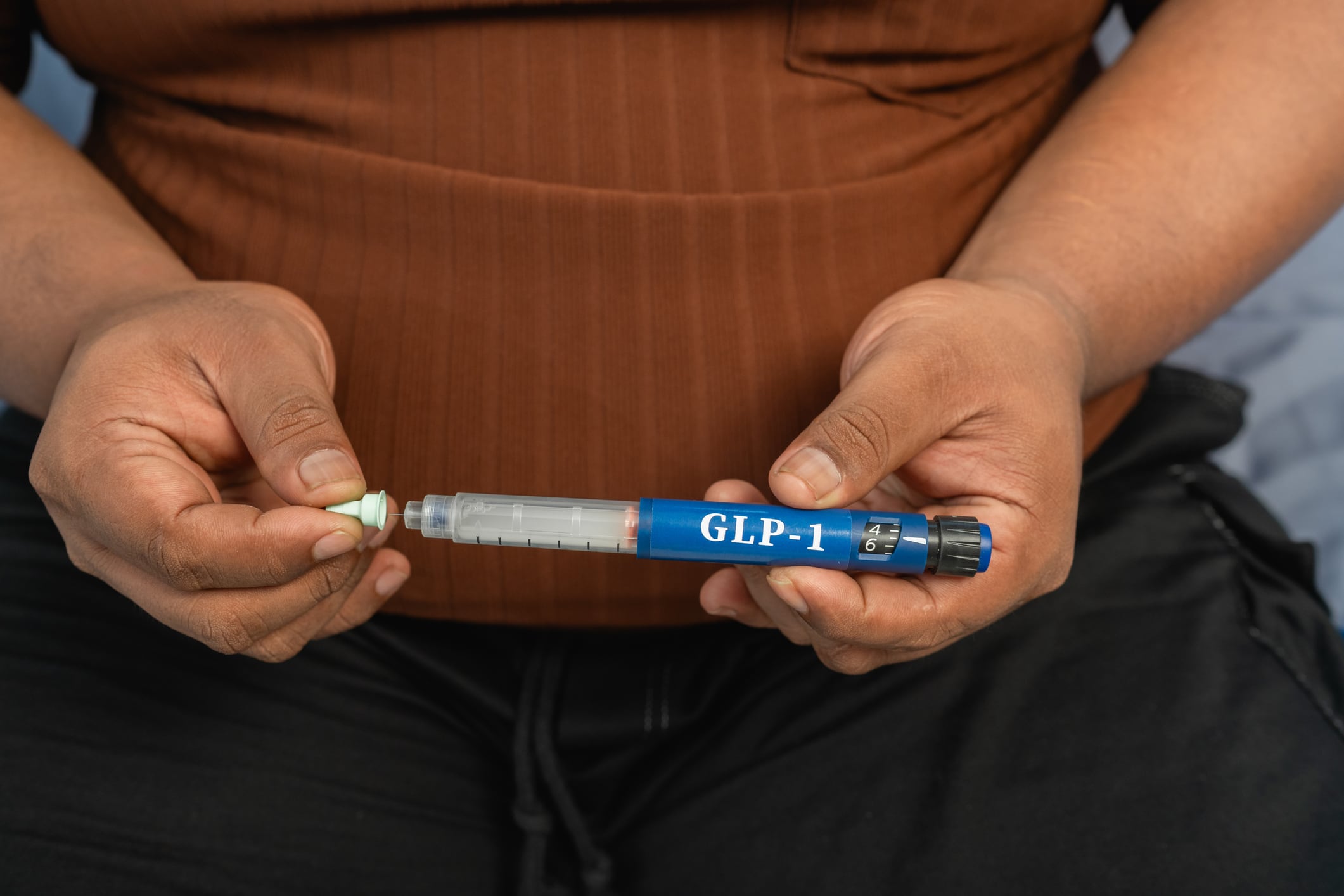 Close-up view of an obese man's hands attaching a needle or adjusting the dosage mechanism on a blue generic GLP-1 subcutaneous injection pen.