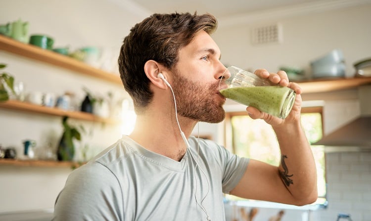 One fit young caucasian man drinking a glass of healthy green detox smoothie while wearing earphones in a kitchen at home. Guy having fresh fruit juice to cleanse and provide energy for training. Wholesome drink with vitamins and nutrients