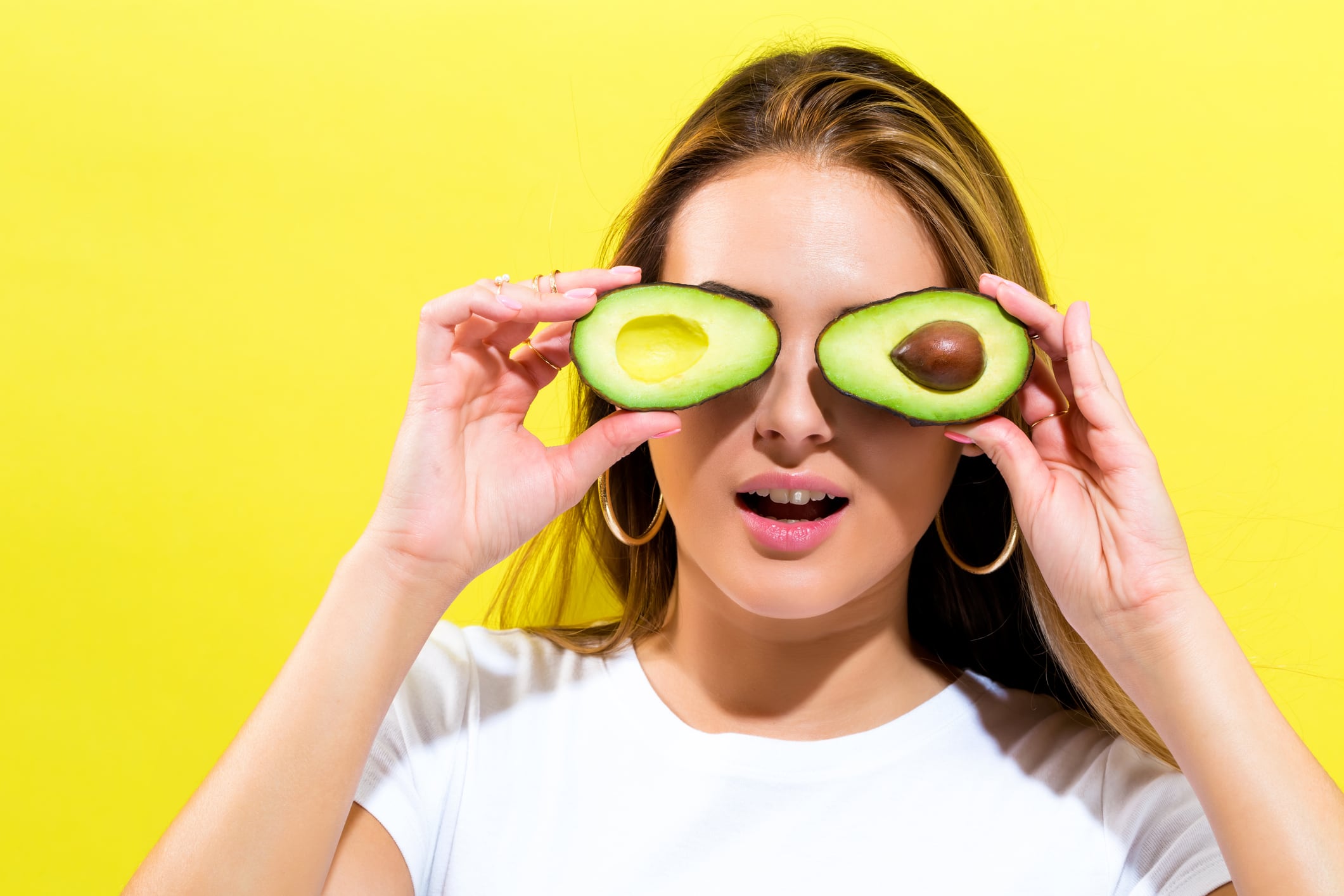 Happy young woman holding avocado halves on a yellow background