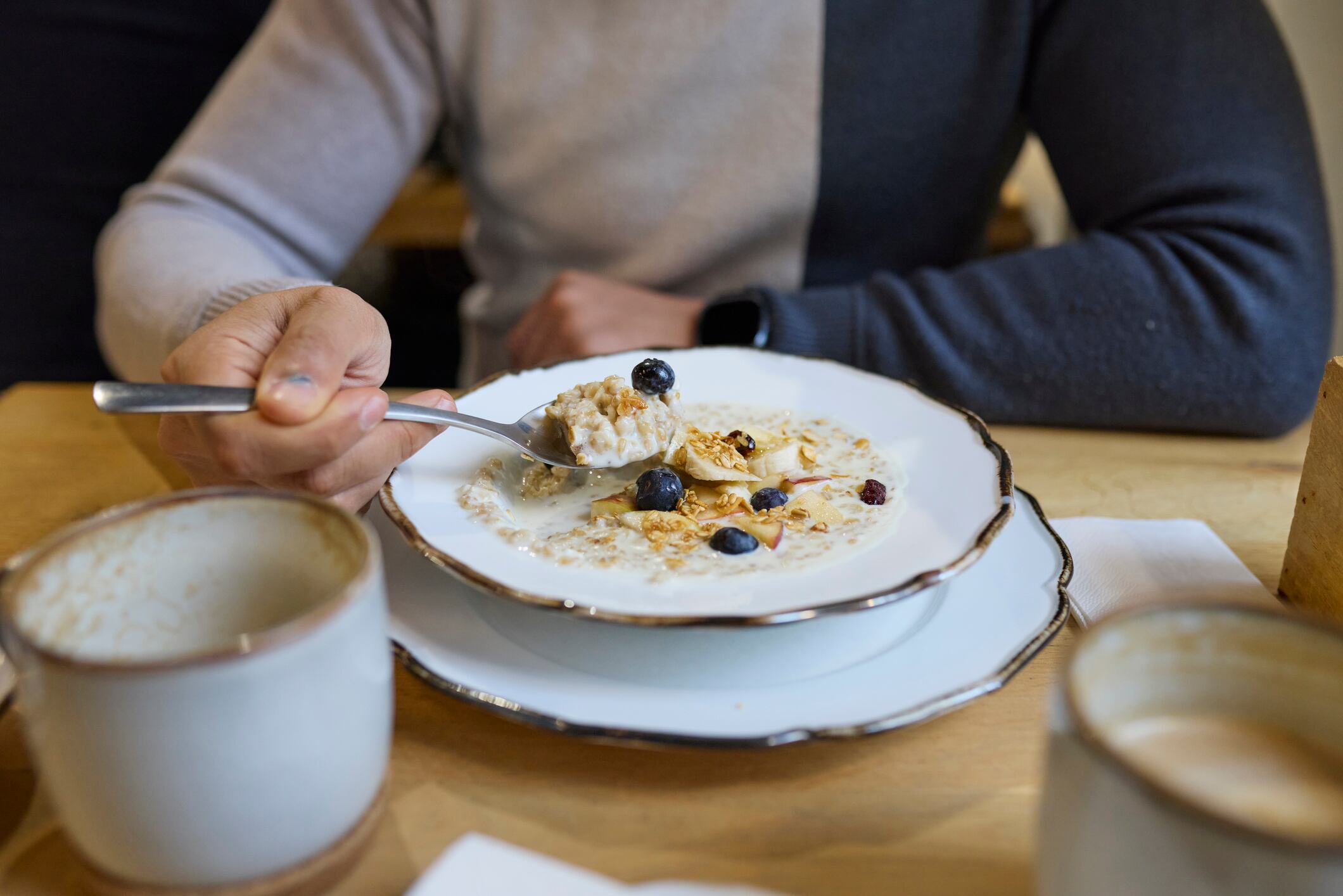 Overhead wide view of man eating oat pudding topped with apple, blueberries and mixed nuts at a café table. Hands holding a spoon over a breakfast plate, with coffee cups on a wooden table, showing healthy morning meal, Food and Drink, Food, casual dining and everyday café lifestyle indoors.