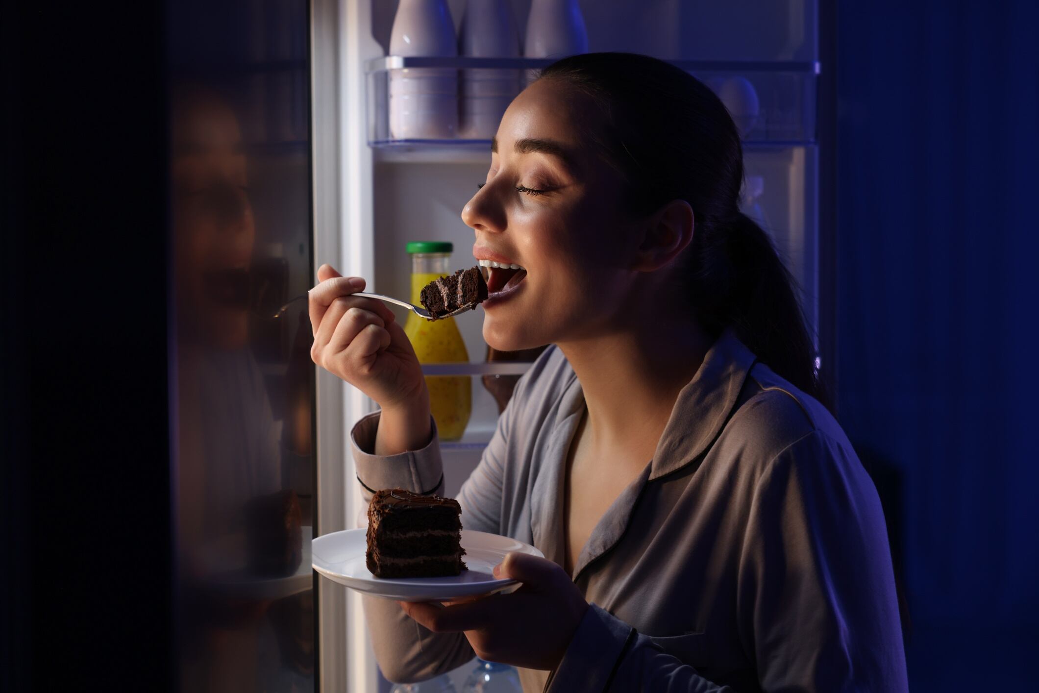 Young woman eating cake near refrigerator in kitchen at night.