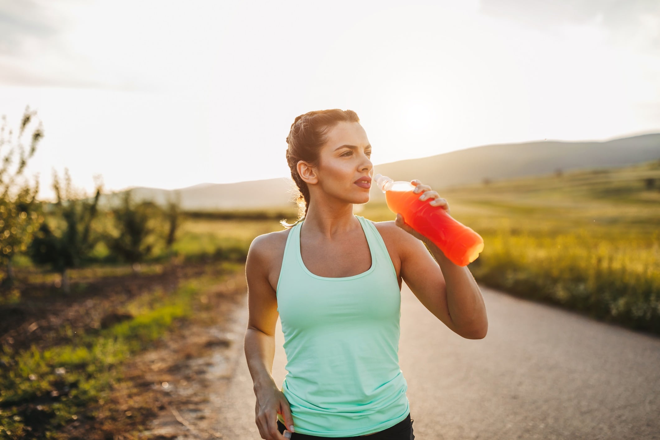 Attractive young blonde sportswoman is refreshing with cold drink after jogging in nature on sunny summer day at sunset