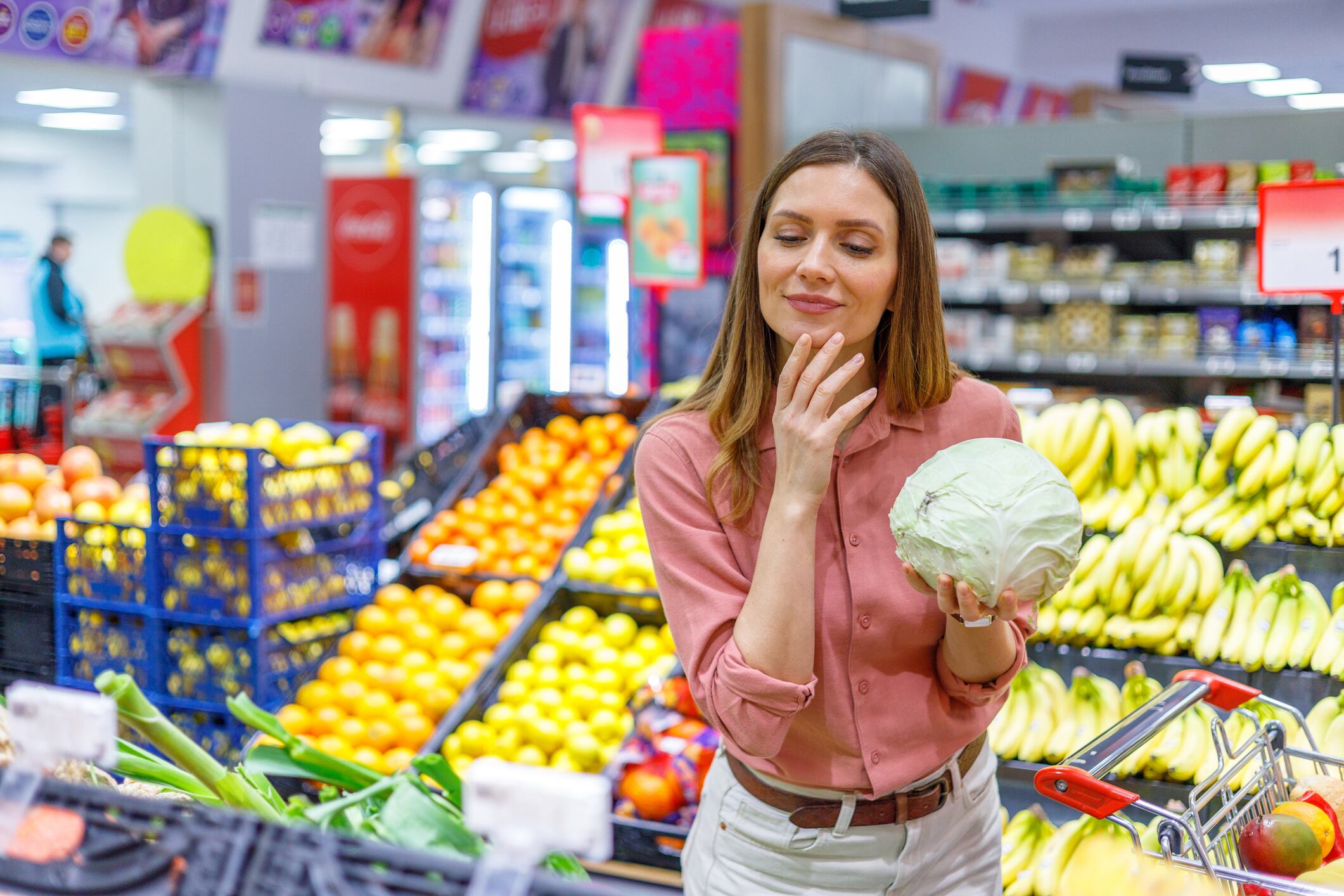 Woman standing in a supermarket produce aisle, holding a head of cabbage and contemplating her healthy food choices with a shopping cart nearby
