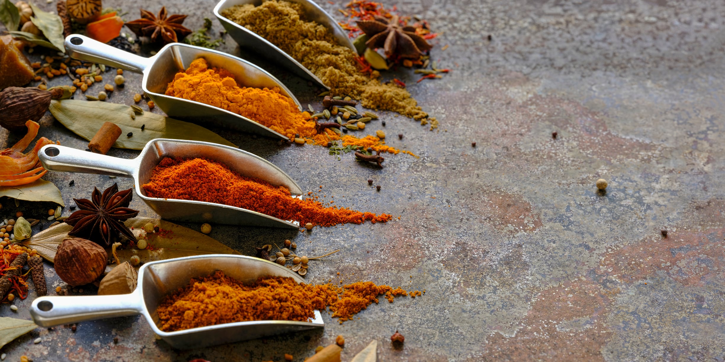 Many colorful, organic, dried, vibrant Indian food, ingredient spices in small aluminum metal spice scoops are arranged in a line on an old, weathered, rusted metal plate background. Taken from a high angle.
