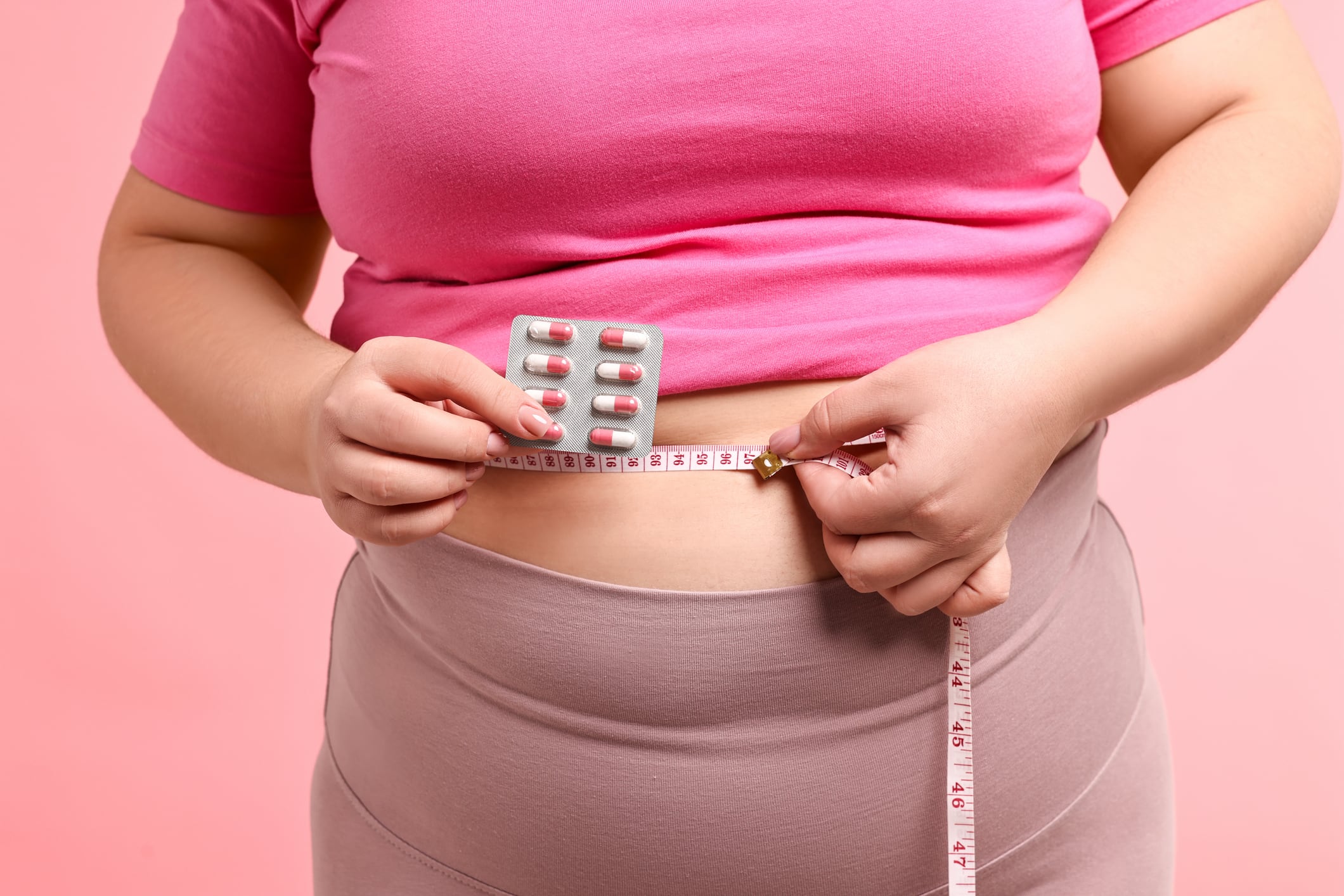 Plus size woman measuring waist with tape and holding blister of weight loss supplements on pink background, closeup