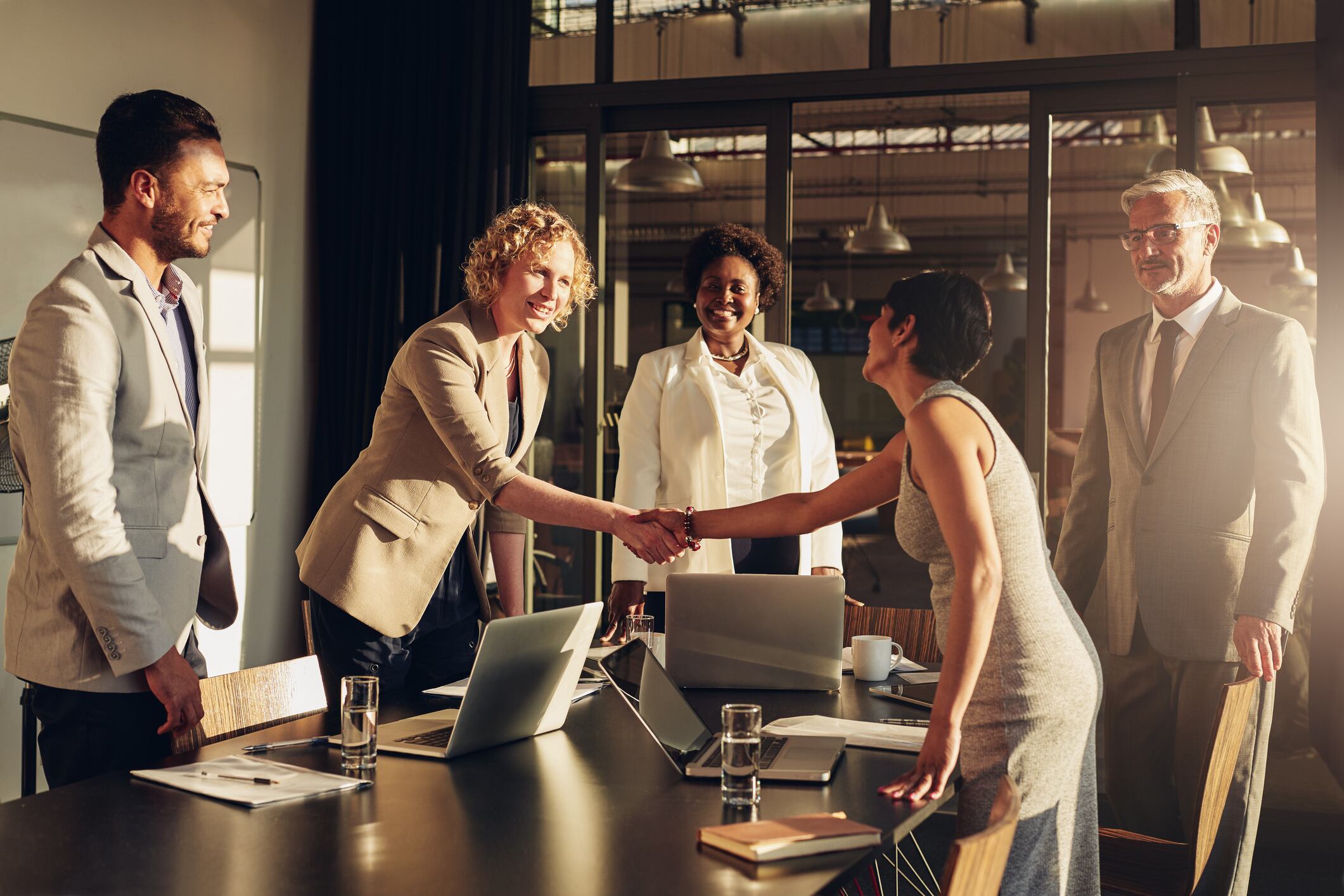 Two businesswomen shaking hands together over a table during a meeting with colleagues in a boardroom in the late afternoon.